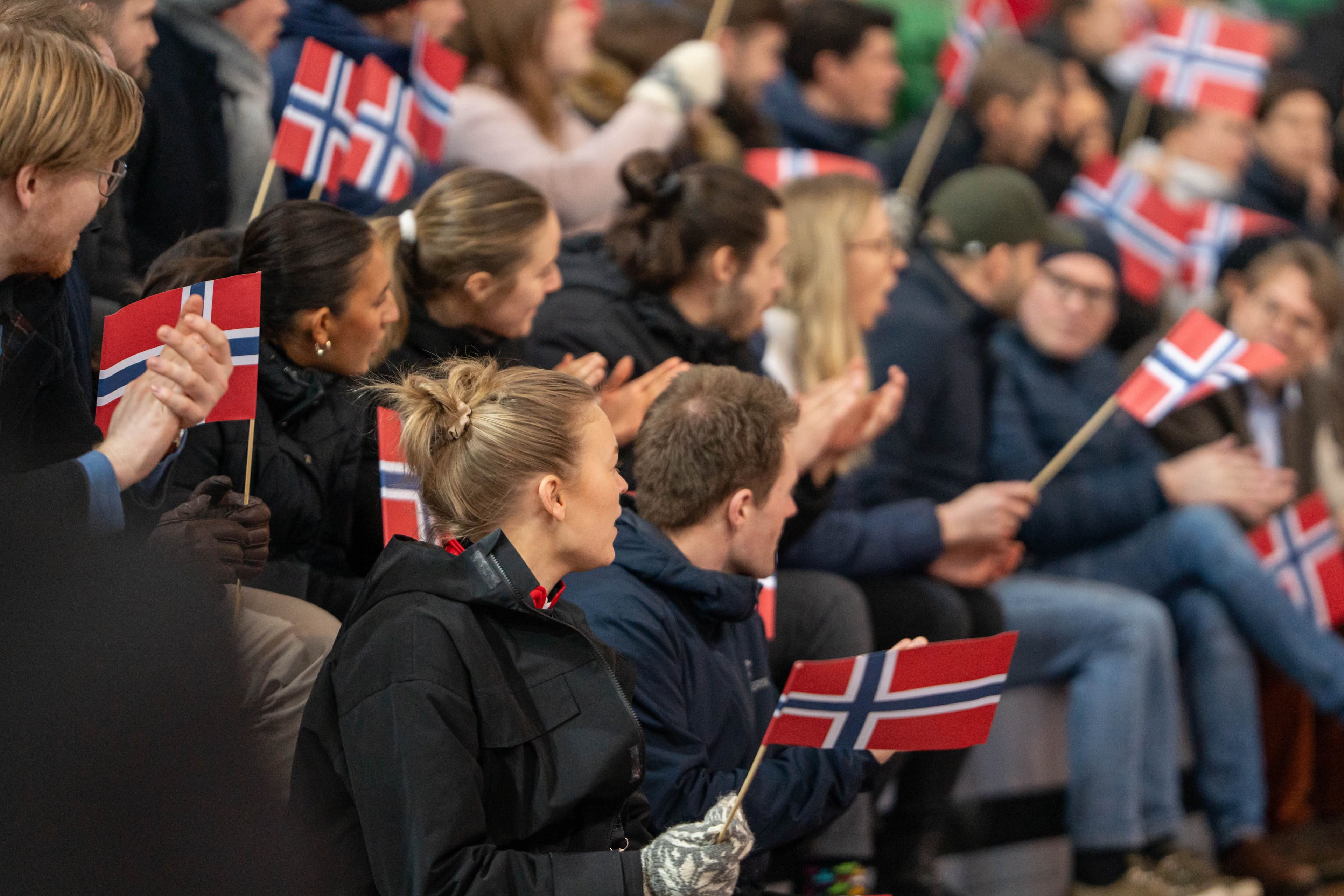 Publikum på en tribune vinker med små norske flagg og applauderer.