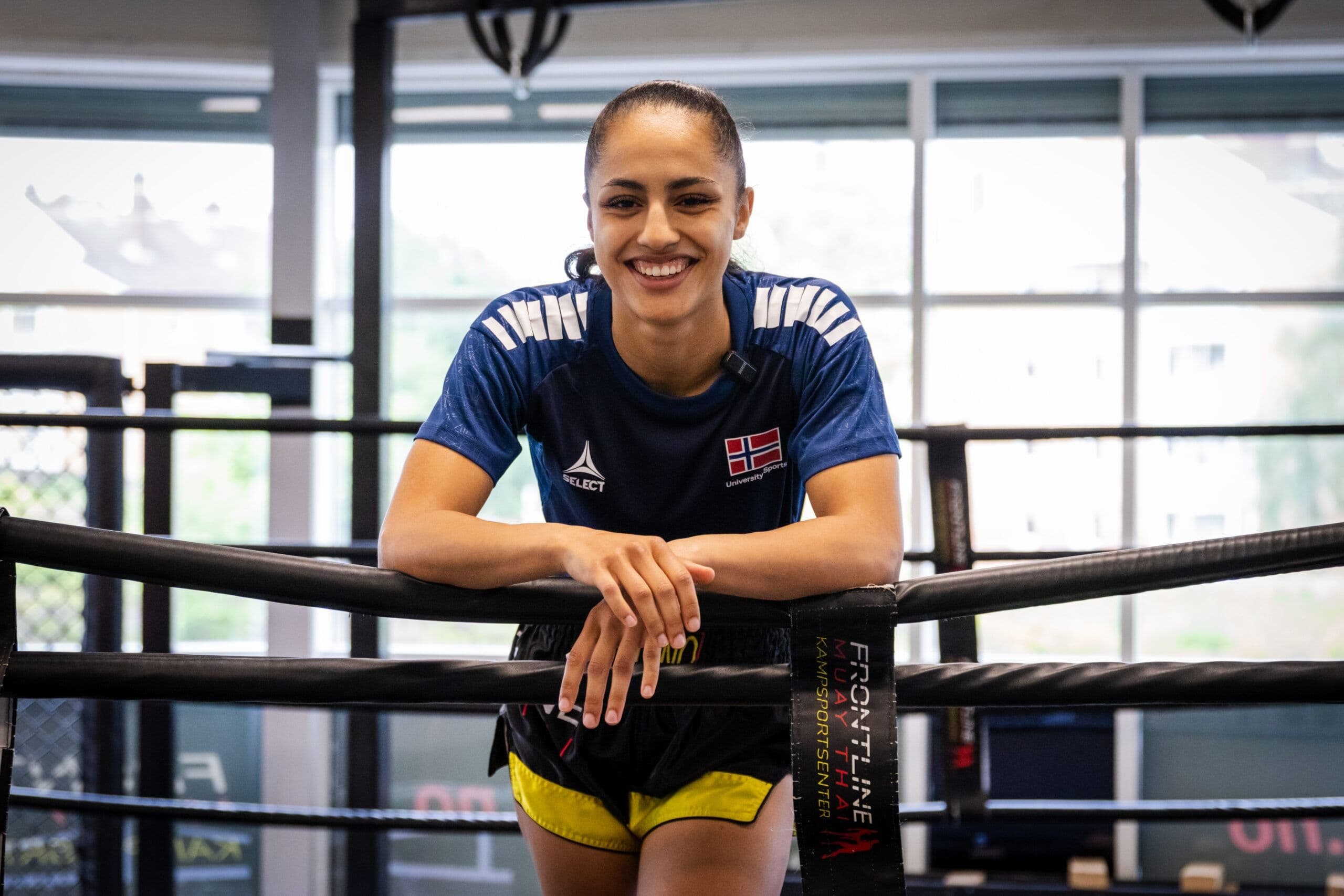 En smilende kvinne i blå «University Sports»-t-skjorte lener seg mot tauene i en muay thai-ring.