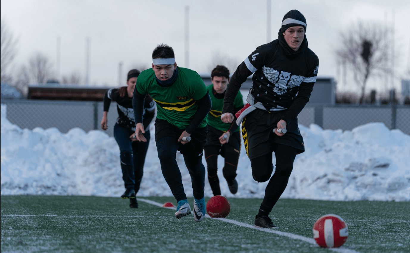 Spillere med kosteskaft løper etter baller på en snødekt kunstgressbane under en rumpeldunk-kamp.