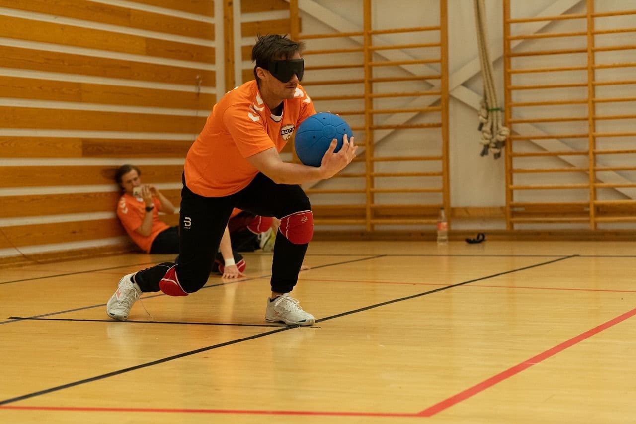 En utøver med øyemaske og oransje drakt går ned på kne og holder en blå goalball i en gymsal med ribbevegger.