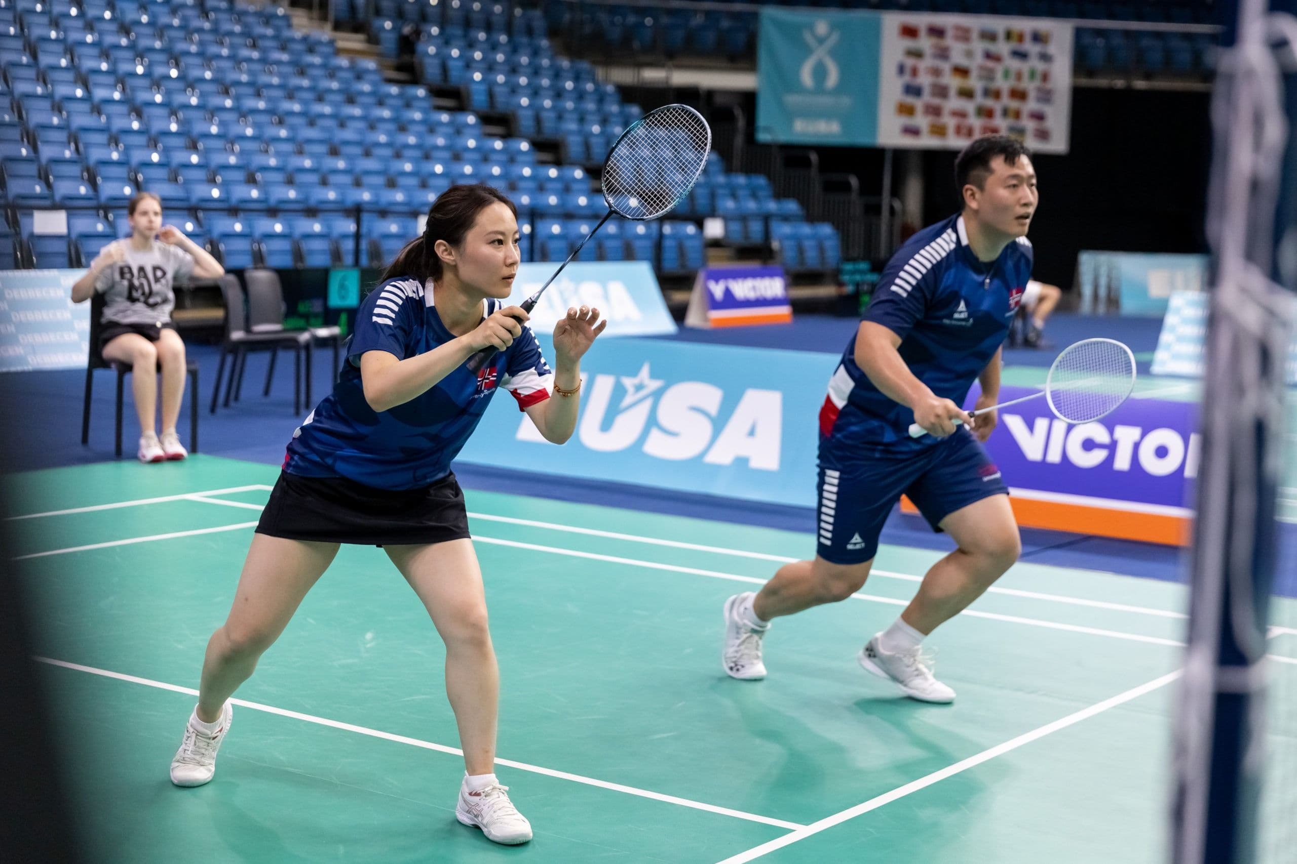 To badmintonspillere i blå drakter står klare på banen under en dobbelkamp.