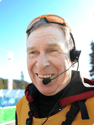 En smilende mann med hodesett og skibriller i oransje jakke ved et skianlegg.