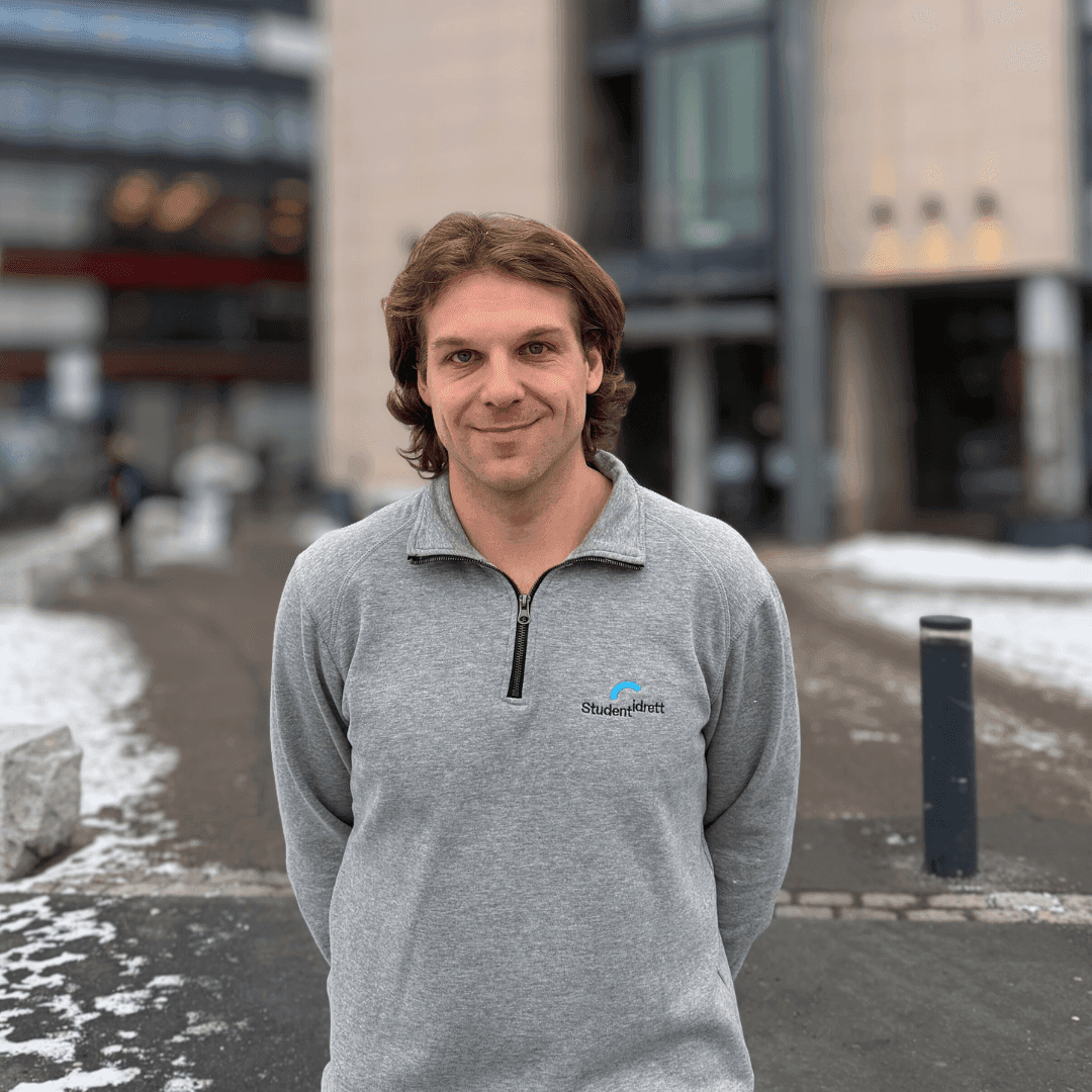 En smilende mann i grå Studentidrett-genser står utenfor en campusbygning med snø.