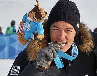 En mann i svart lue biter i en medalje og holder en maskott-bamse, med snø i bakgrunnen.