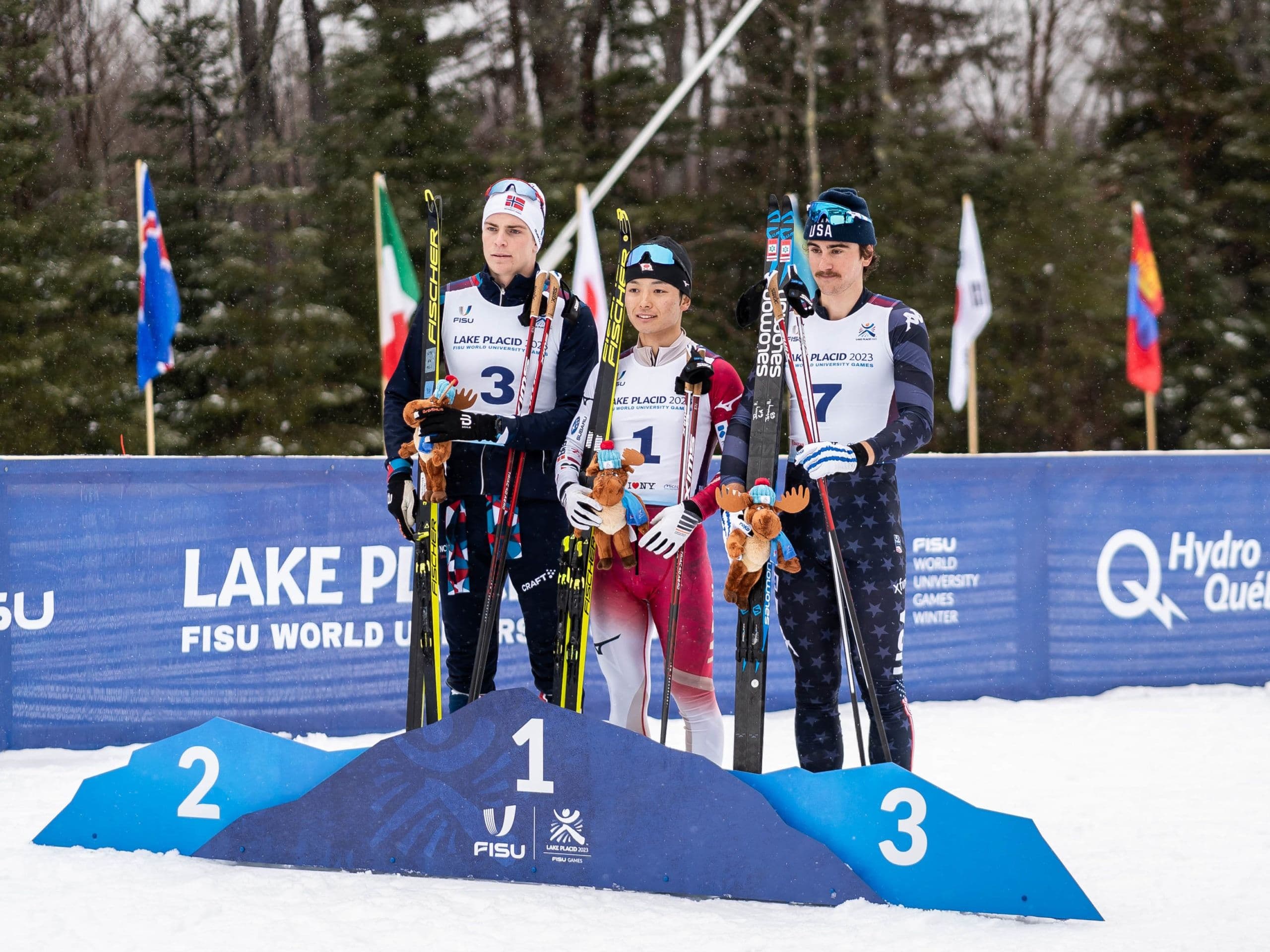 Tre langrennsløpere står på seierspallen med ski og maskotter under FISU World University Games i Lake Placid 2023.