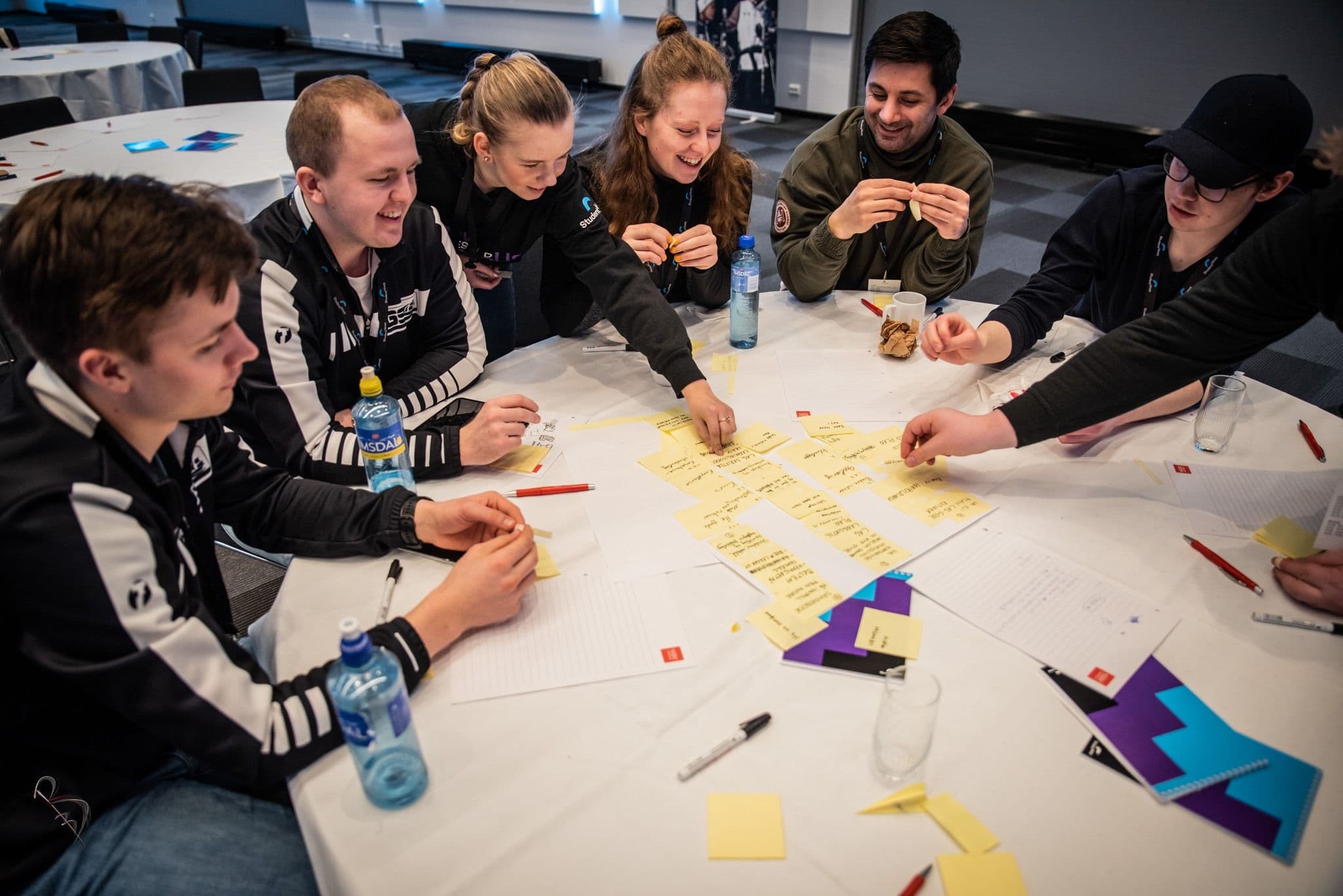 En gruppe unge mennesker jobber med gule lapper og notater rundt et bord under en workshop.