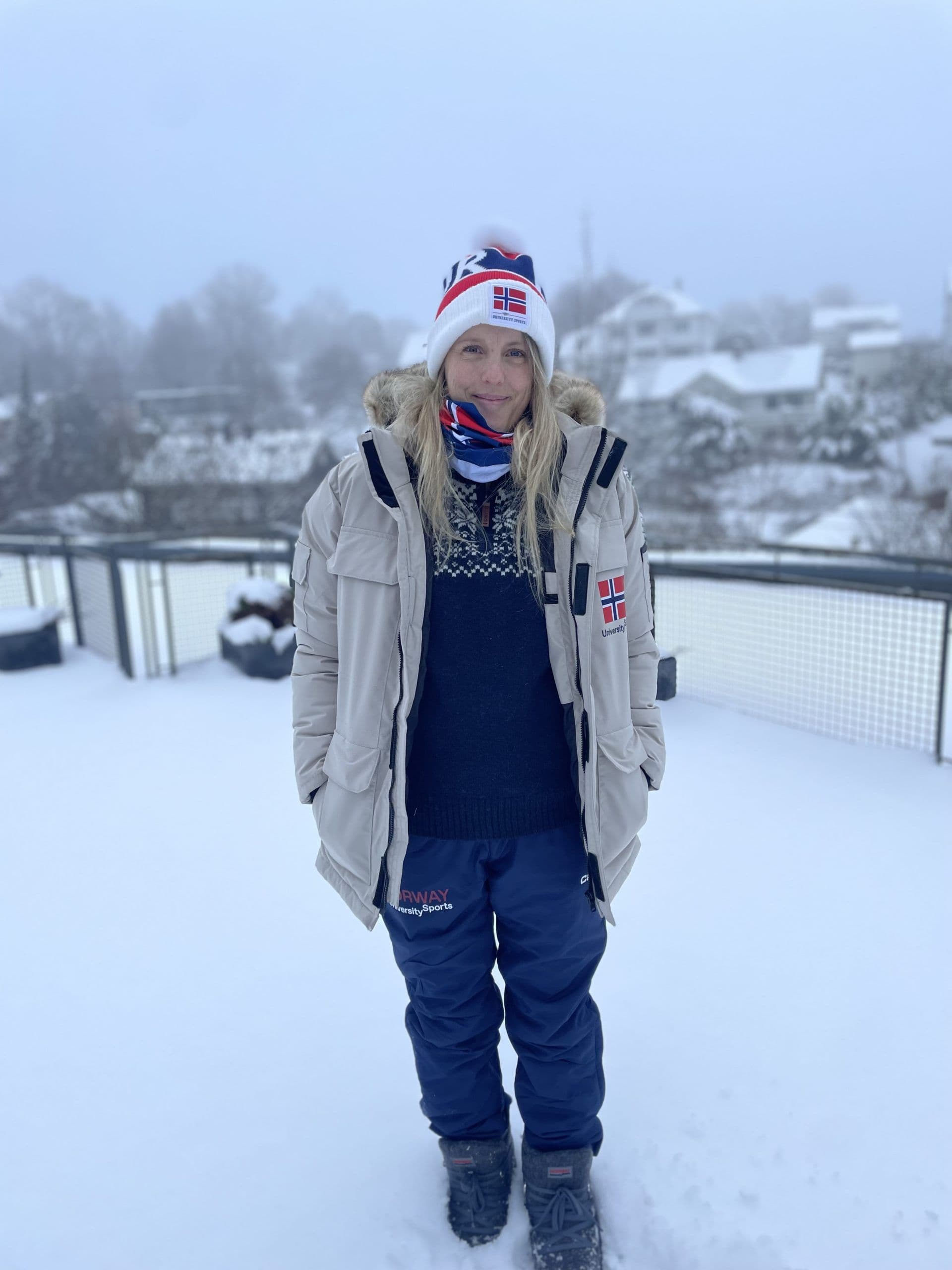 En kvinne i beige parkas og lue med norsk flagg står ute i snøen med snødekte hus i bakgrunnen.
