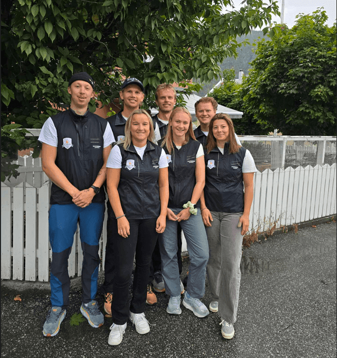En gruppe på sju personer i mørke vester poserer foran et hvitt stakittgjerde.
