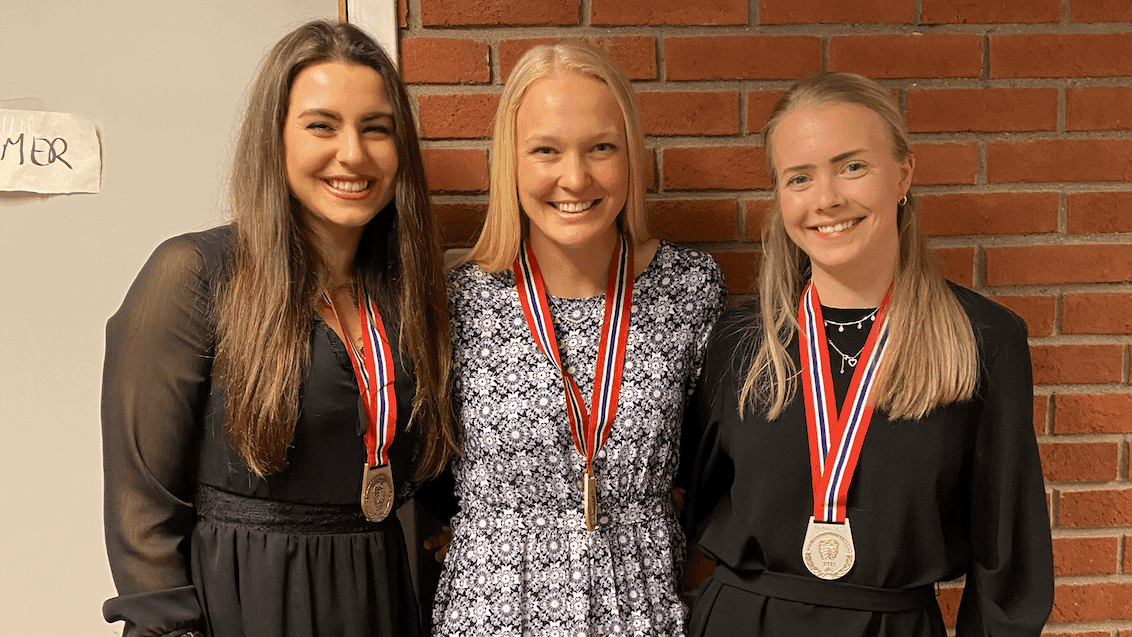 Tre unge kvinner med medaljer rundt halsen smiler foran en teglsteinsvegg.