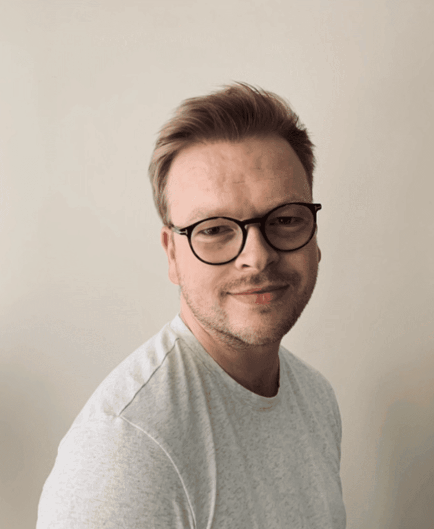 Portrett av en mann med briller i grå t-skjorte mot en lys vegg.