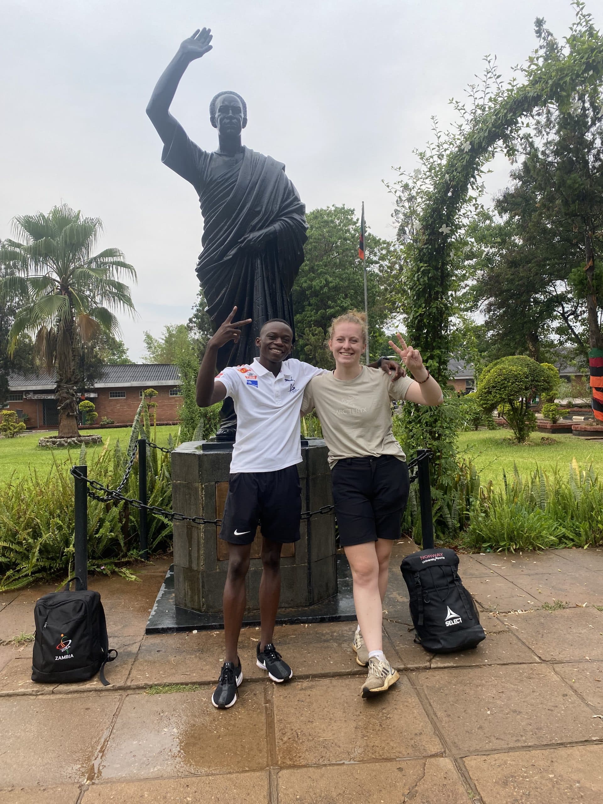 To personer viser seierstegn foran en statue i en park, med ryggsekker merket «Zambia» på bakken.