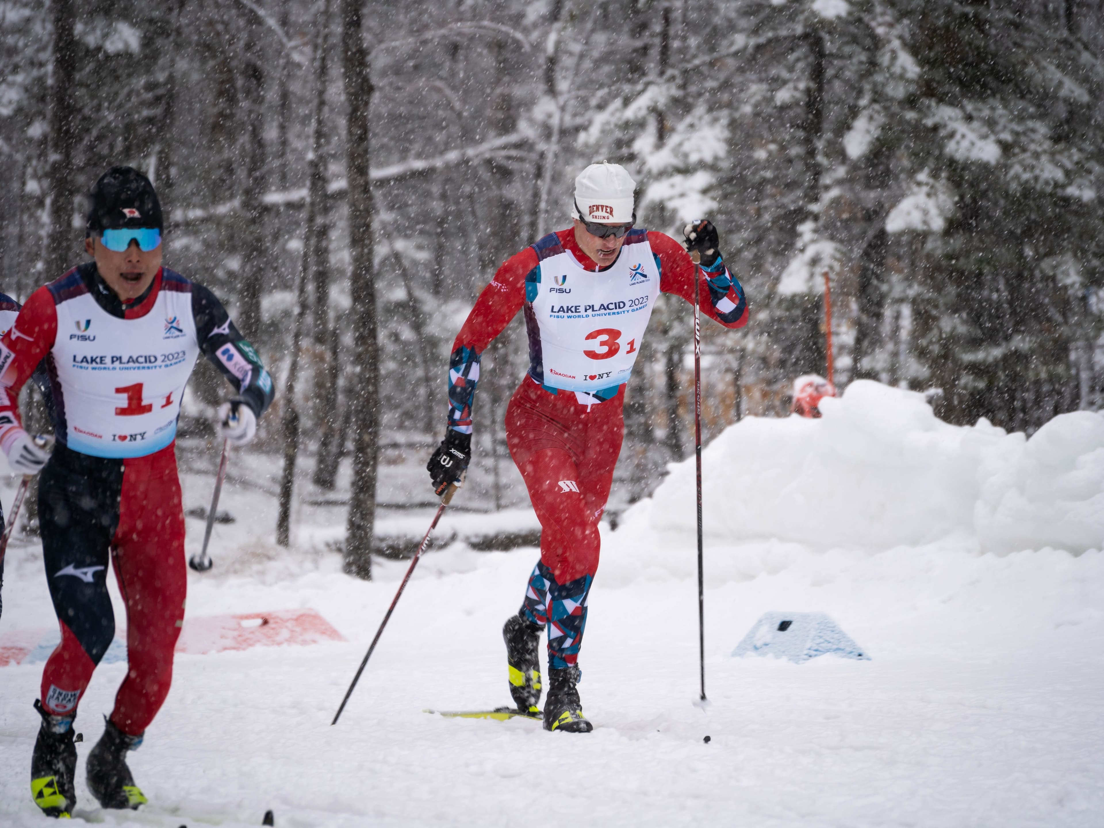 To langrennsløpere i landslagsdrakter går i løypa i snøvær under FISU World University Games i Lake Placid 2023.
