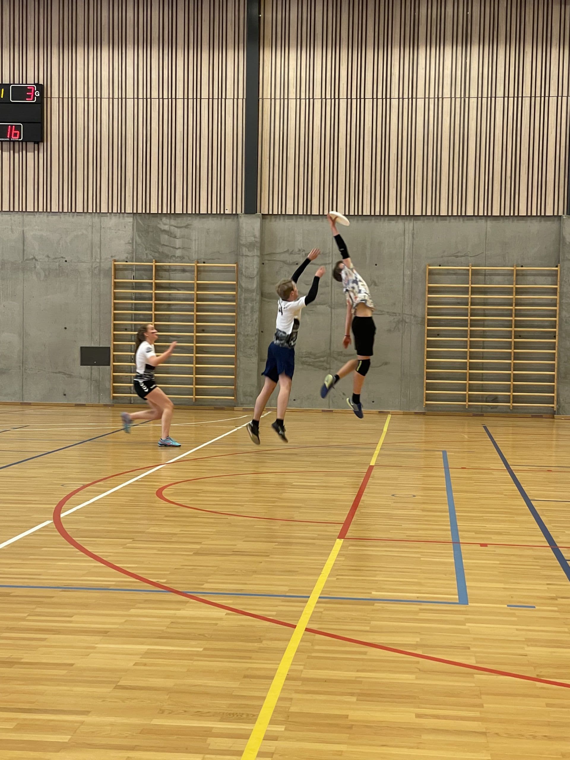 To spillere hopper etter en frisbee i en idrettshall under en ultimate-kamp.