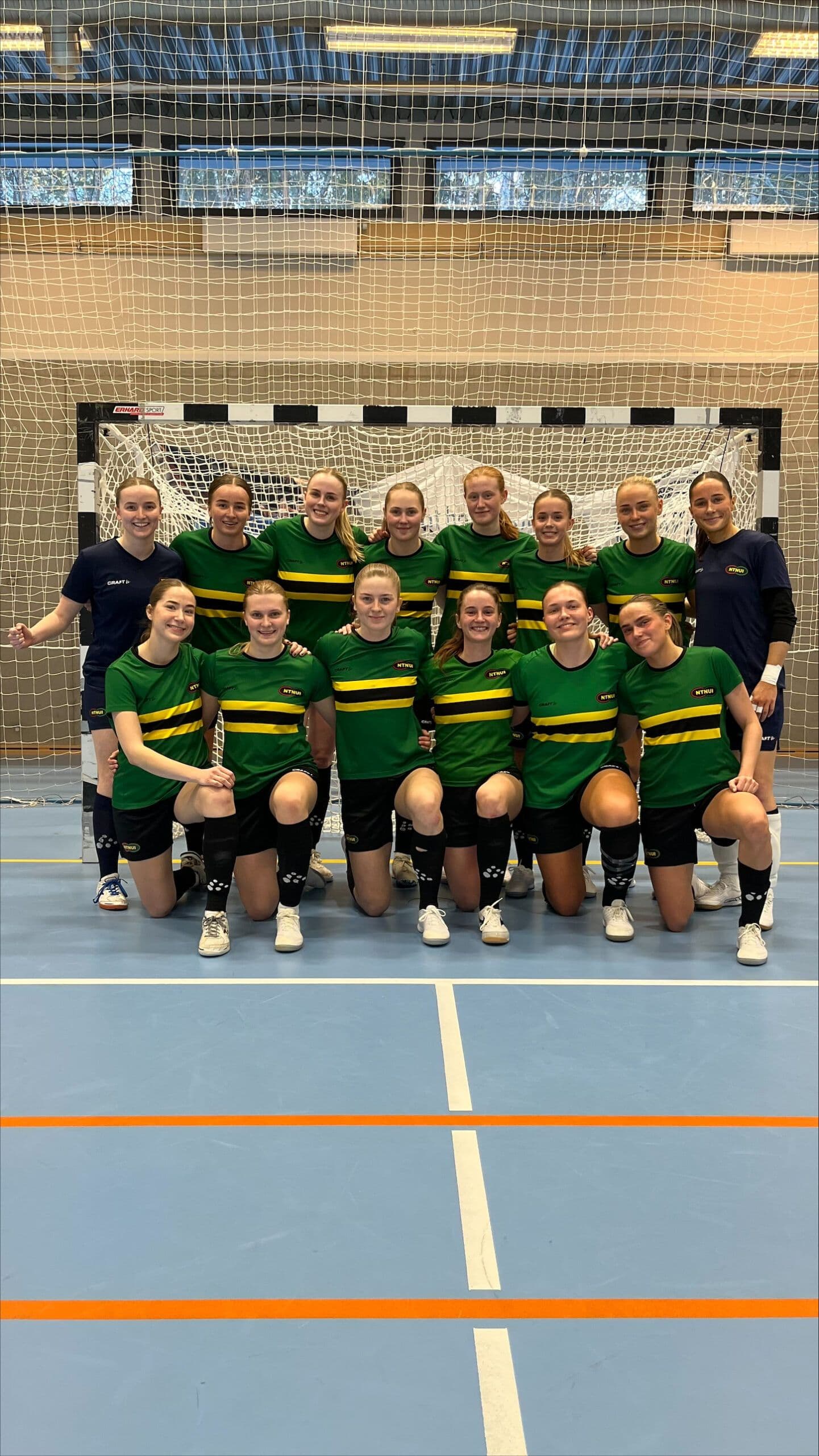 Et kvinnelag i grønne og gule futsaldrakter poserer foran et håndballmål i en idrettshall.
