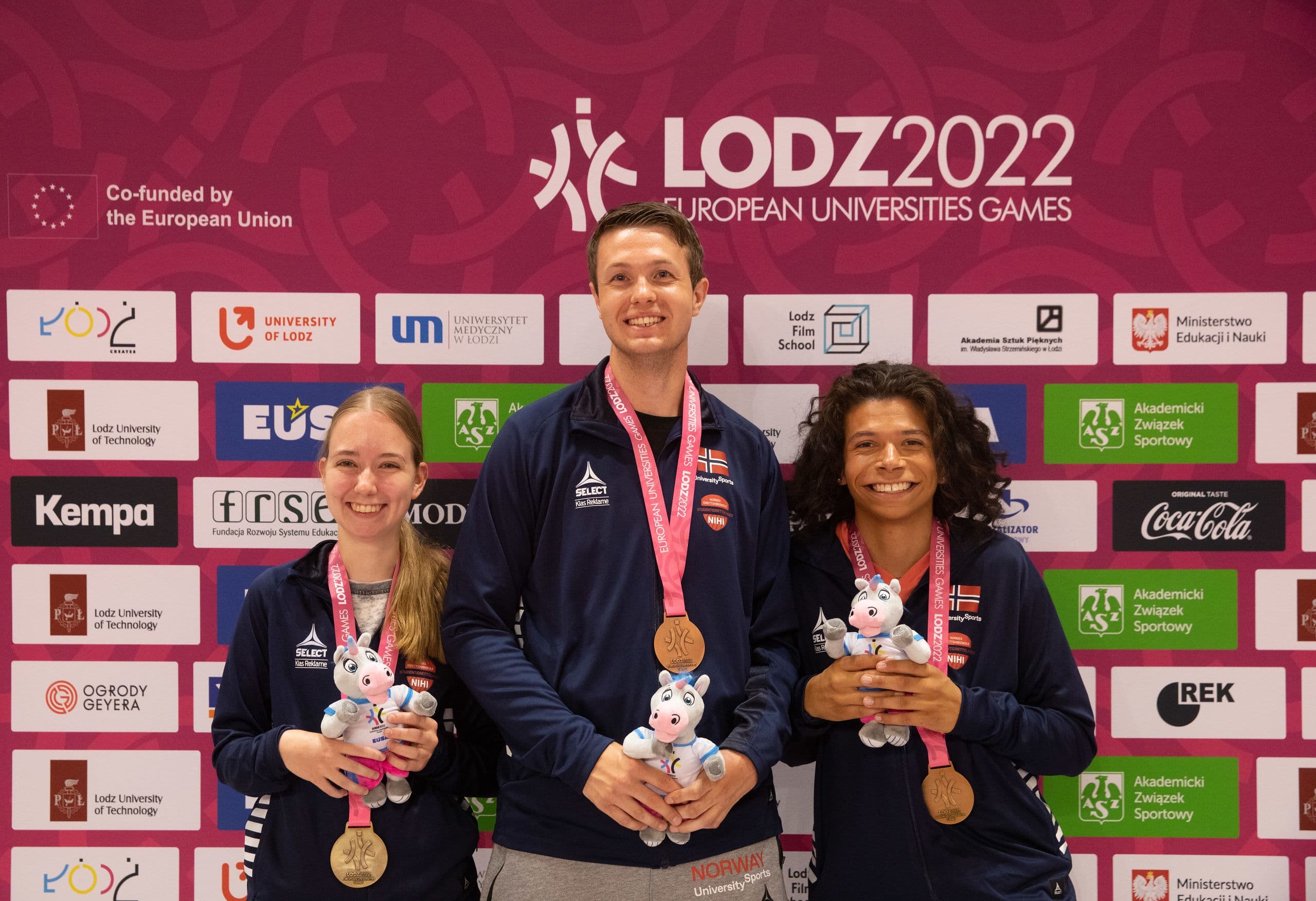 Tre medaljevinnere viser fram medaljer og maskotter foran en vegg med teksten Lodz 2022 European Universities Games.