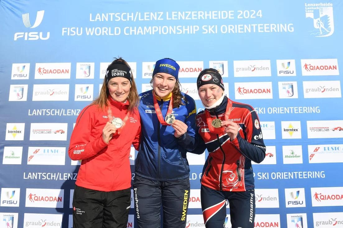 Tre kvinnelige medaljevinnere viser fram medaljene sine på pallen foran en vegg med teksten FISU World Championship Ski Orienteering.