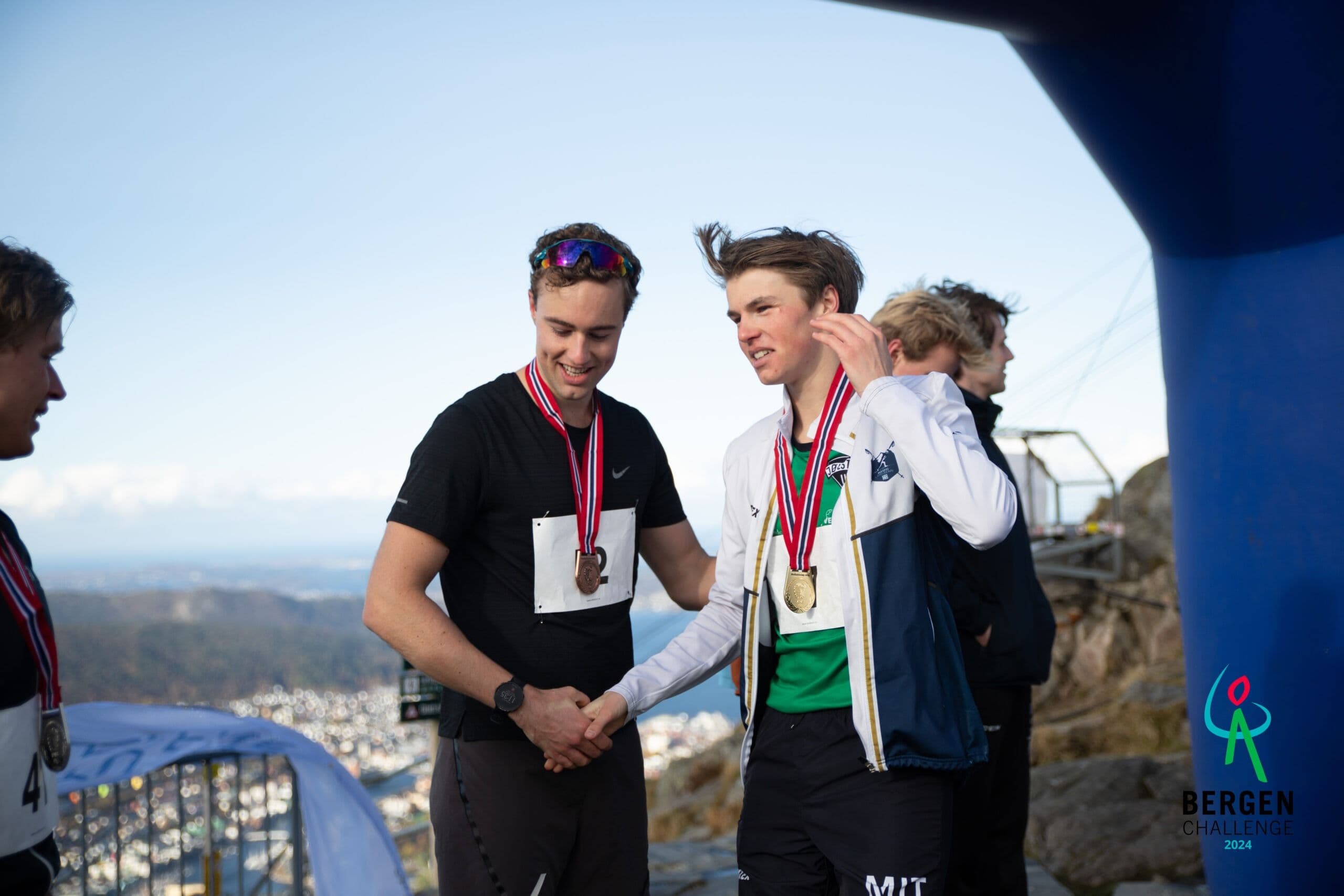 To unge menn med medaljer håndhilser på en fjelltopp med utsikt over Bergen under Bergen Challenge.