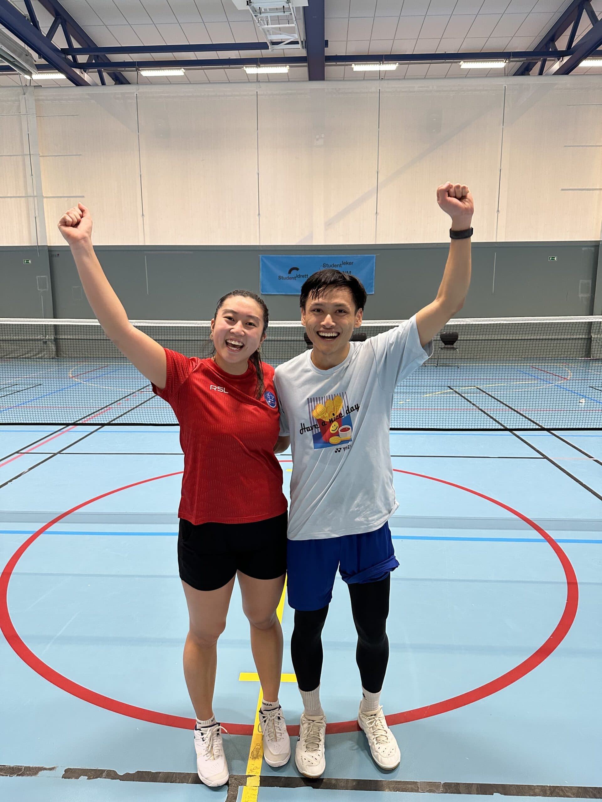 En kvinne og en mann jubler med hevede armer på en badmintonbane i en idrettshall.