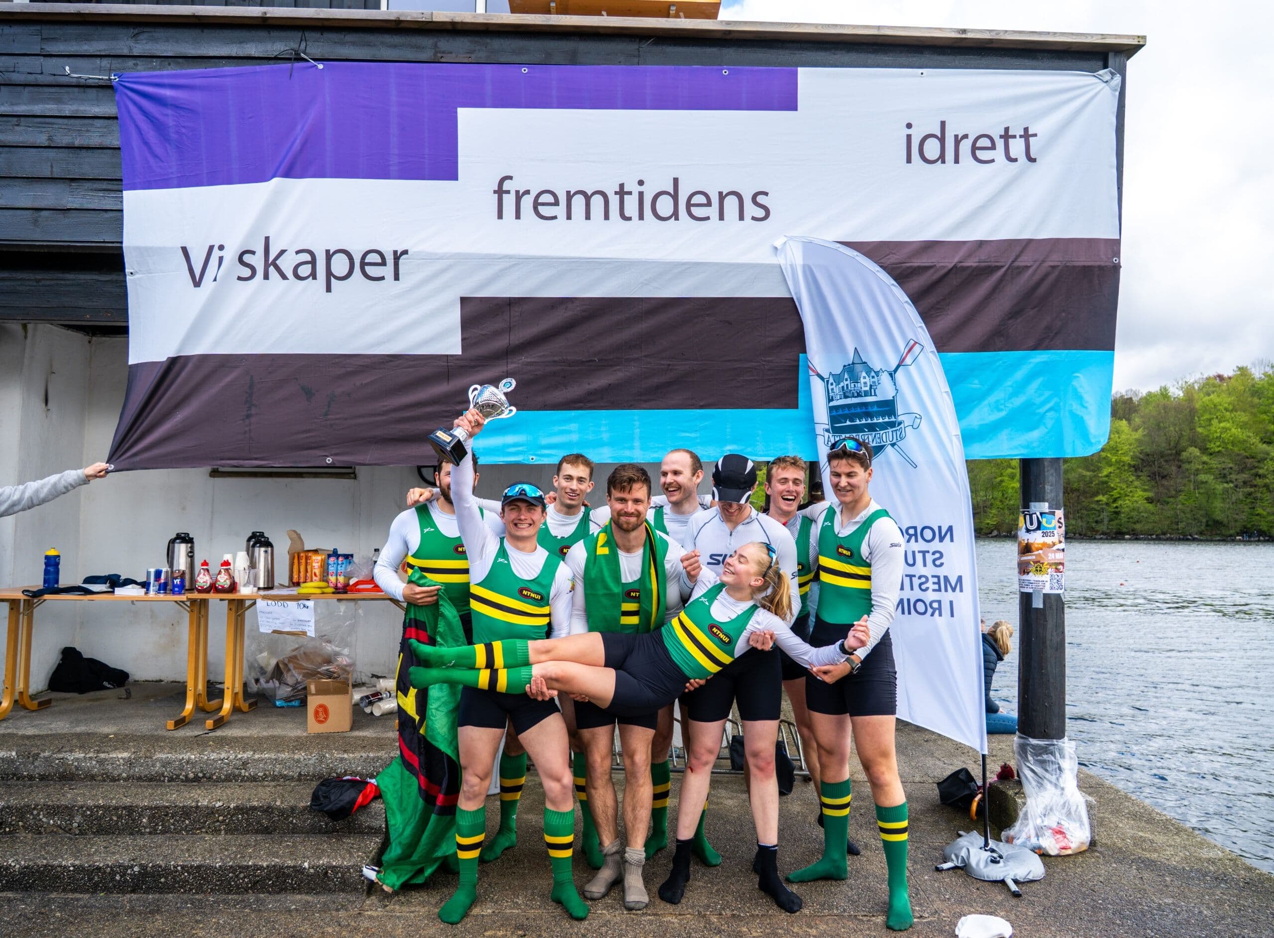 Et jublende roerlag med pokal poserer i grønne og gule drakter foran et Studentidrett-banner med teksten Vi skaper fremtidens idrett.