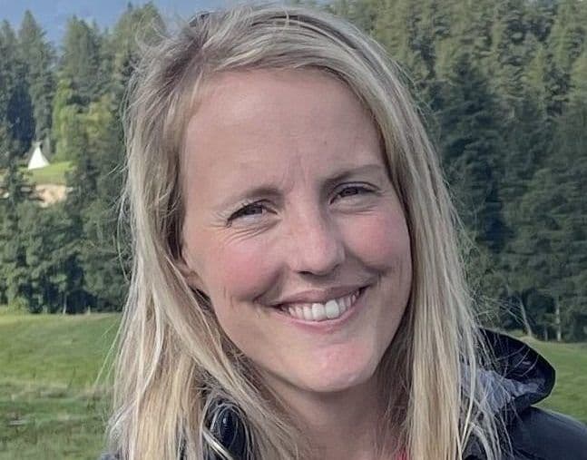 Portrett av en smilende kvinne med lyst hår utendørs foran skog.