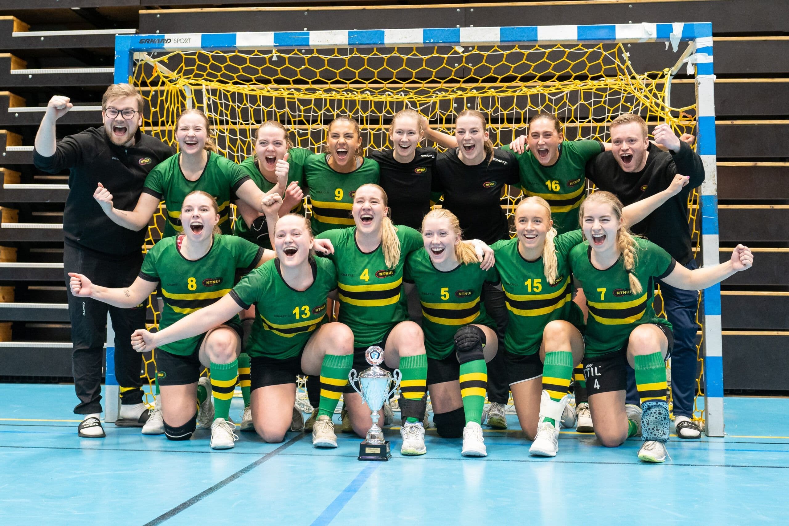 Et jublende kvinnelag i grønne og gule drakter feirer med pokal foran et håndballmål i en idrettshall.
