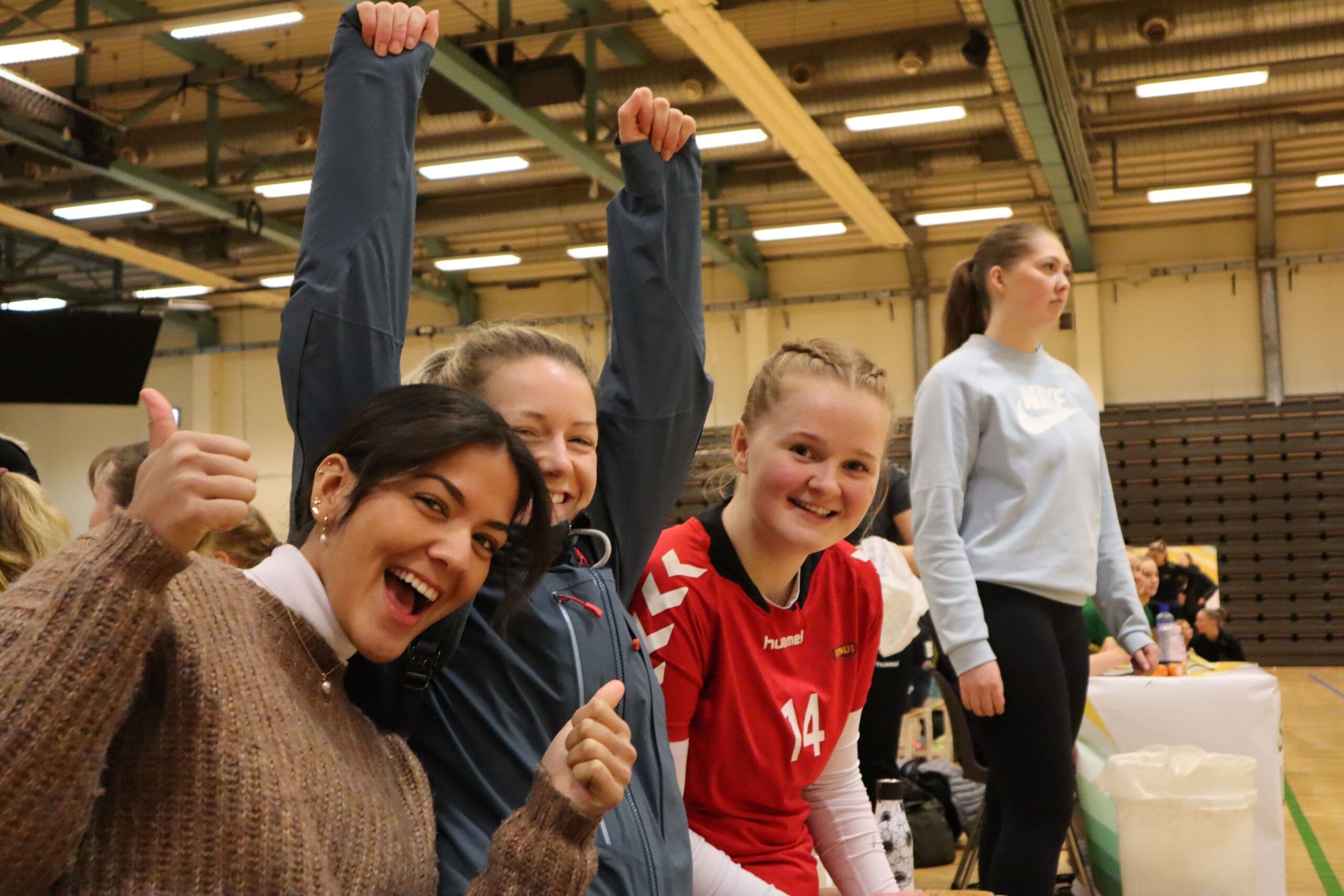 Jublende studenter med tommel opp i en idrettshall, en av dem i rød håndballdrakt med nummer 14.