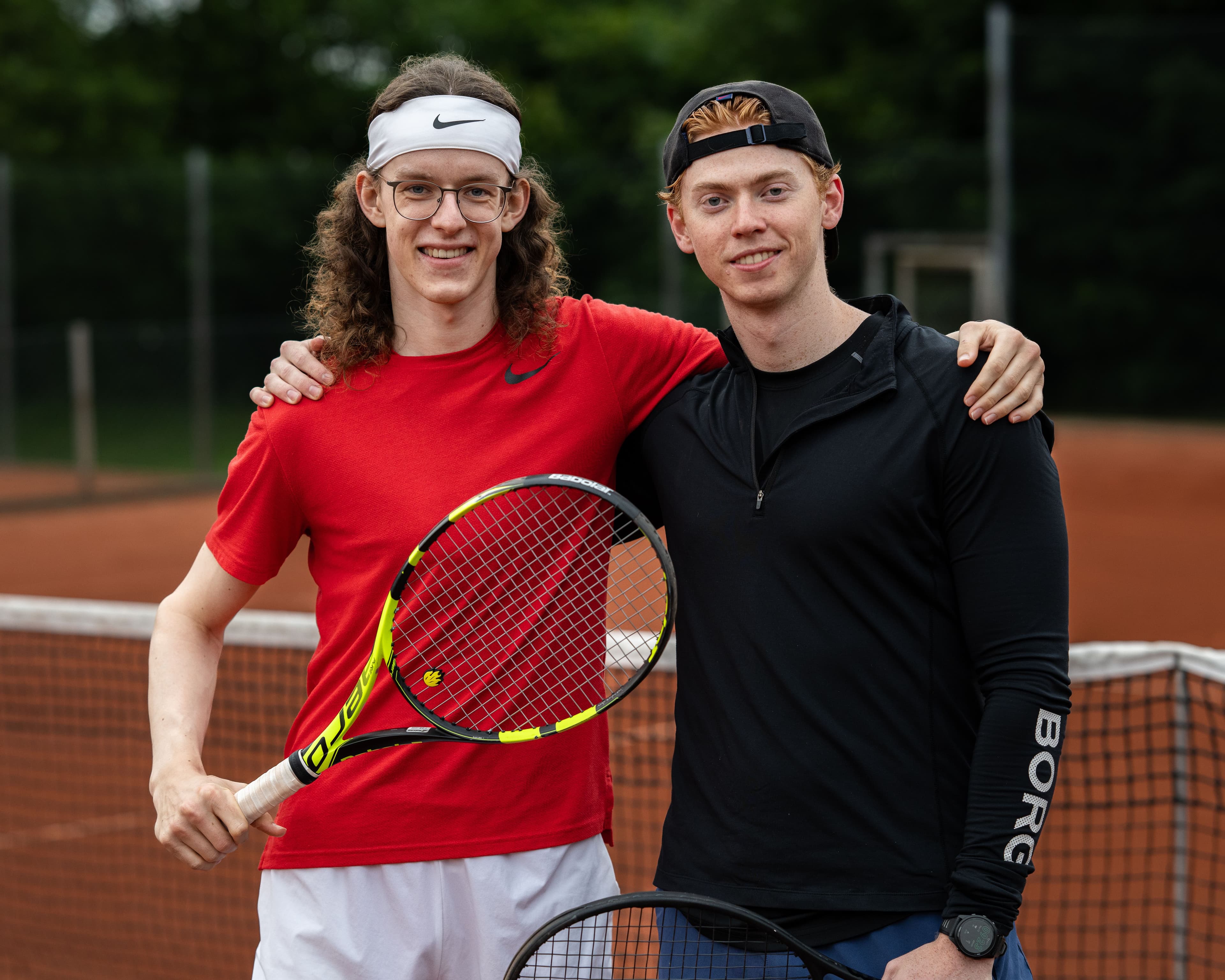 To tennisspillere med racketer står med armene om hverandre ved nettet på en grusbane.