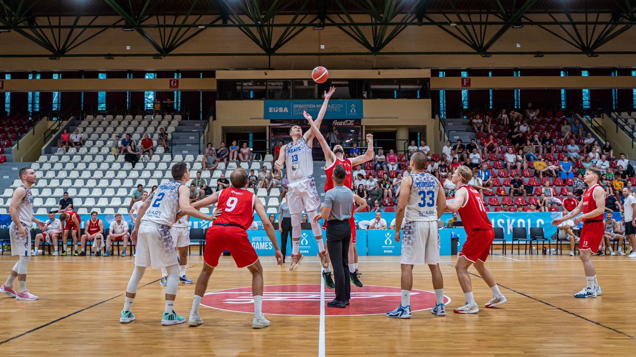 Basketkamp der to spillere hopper etter ballen i tipp-off, med dommer og medspillere rundt på banen.