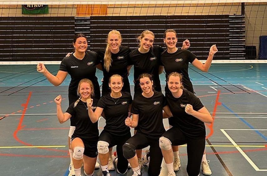 Et kvinnelag i svarte drakter poserer smilende foran nettet i en idrettshall.