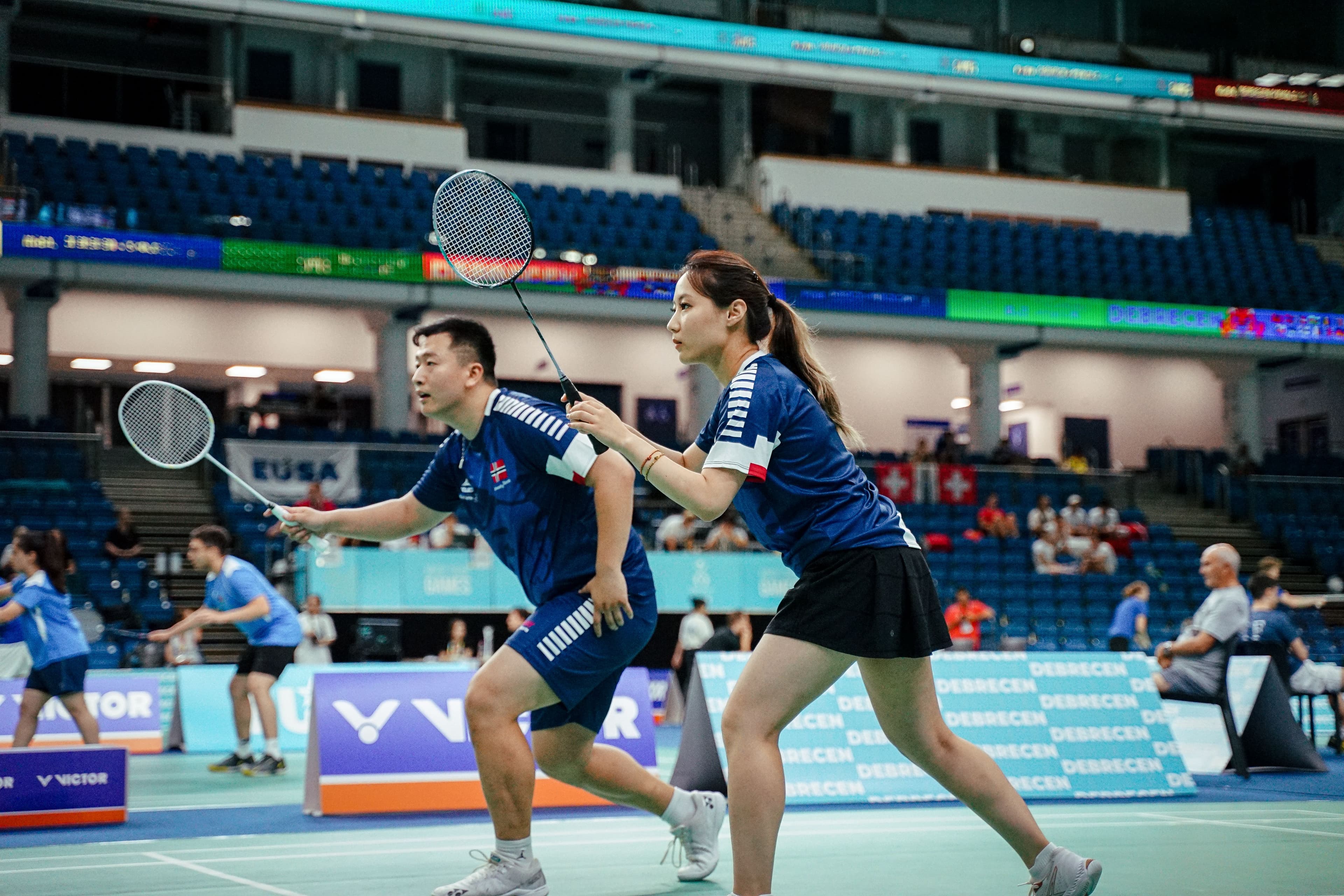 Et badmintonpar i blå drakter spiller double på en innendørs bane.