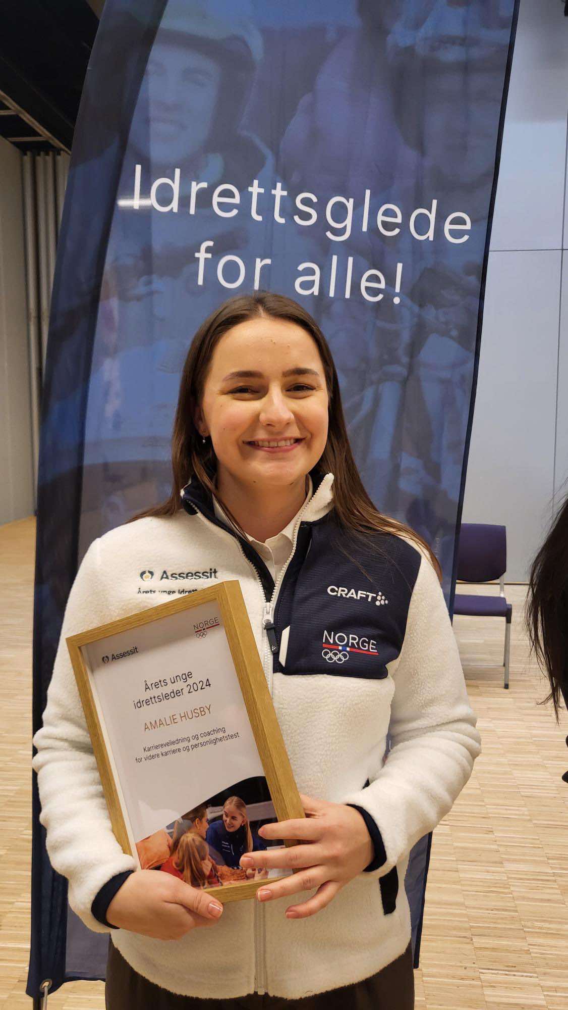 En kvinne holder en innrammet utmerkelse med teksten Årets unge idrettsleder 2024 foran en banner med teksten Idrettsglede for alle!