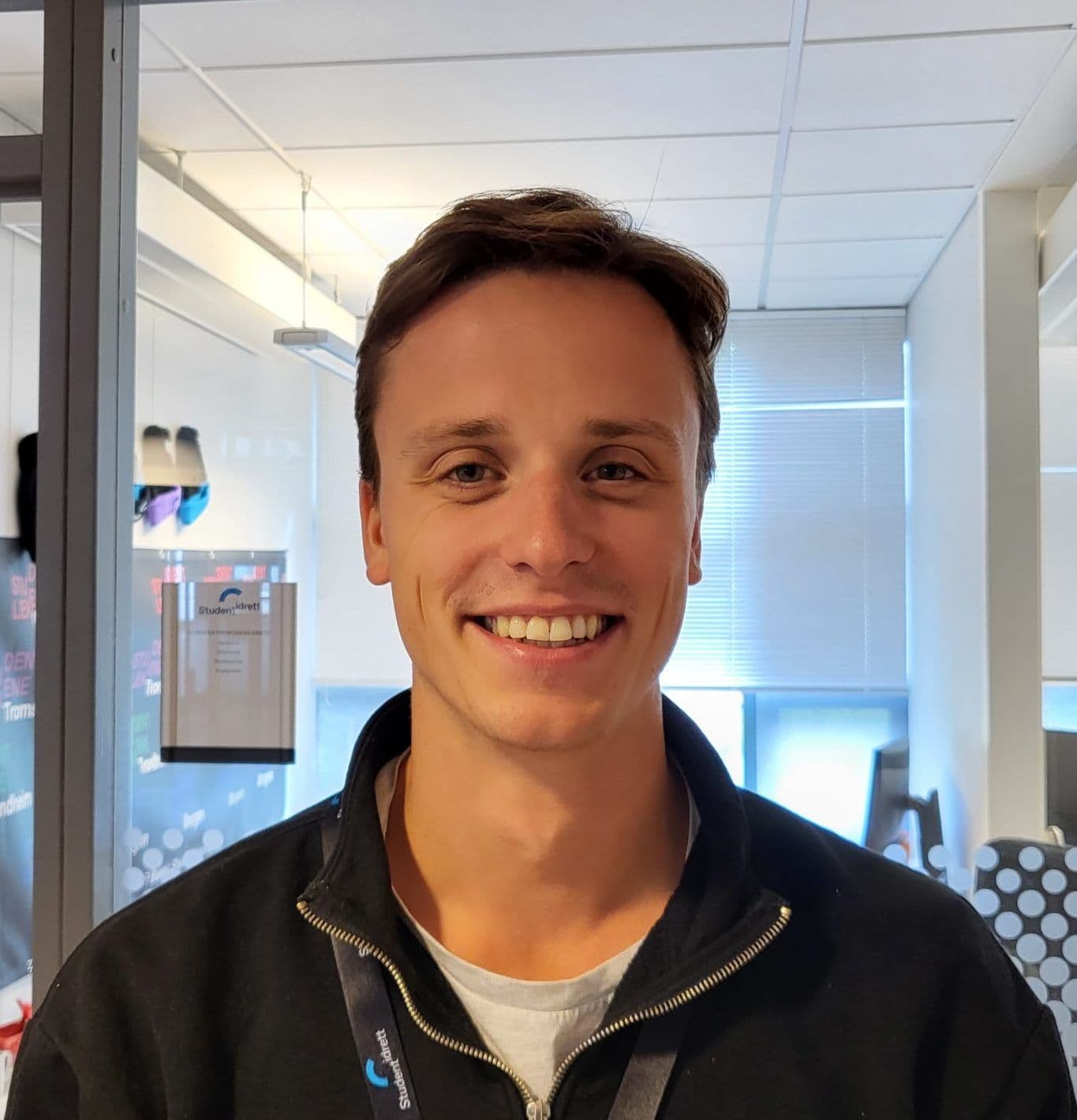 Portrett av en smilende mann i mørk glidelåsgenser i et kontorlandskap.