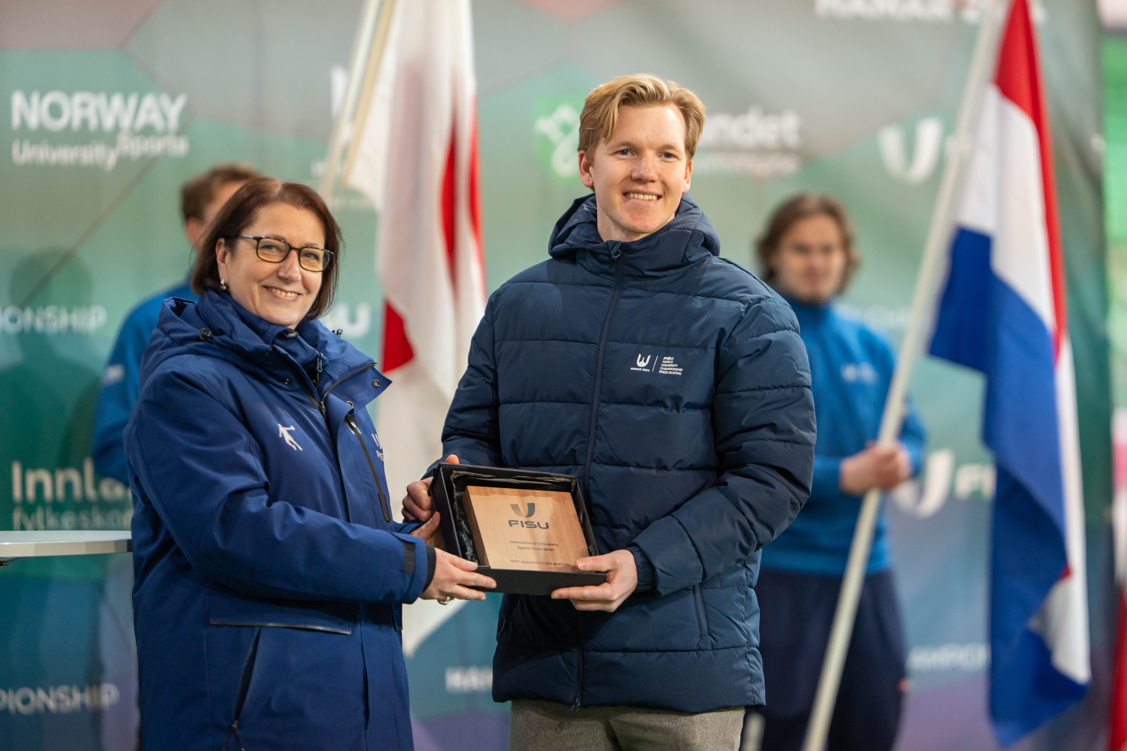 En kvinne overrekker en plakett til en smilende mann i Norway University Sports-jakke foran nasjonsflagg under et FISU-mesterskap.