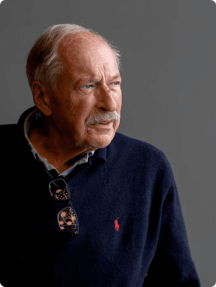 Portrett av en eldre mann i mørkeblå genser med briller hengende i halsen mot en grå bakgrunn.