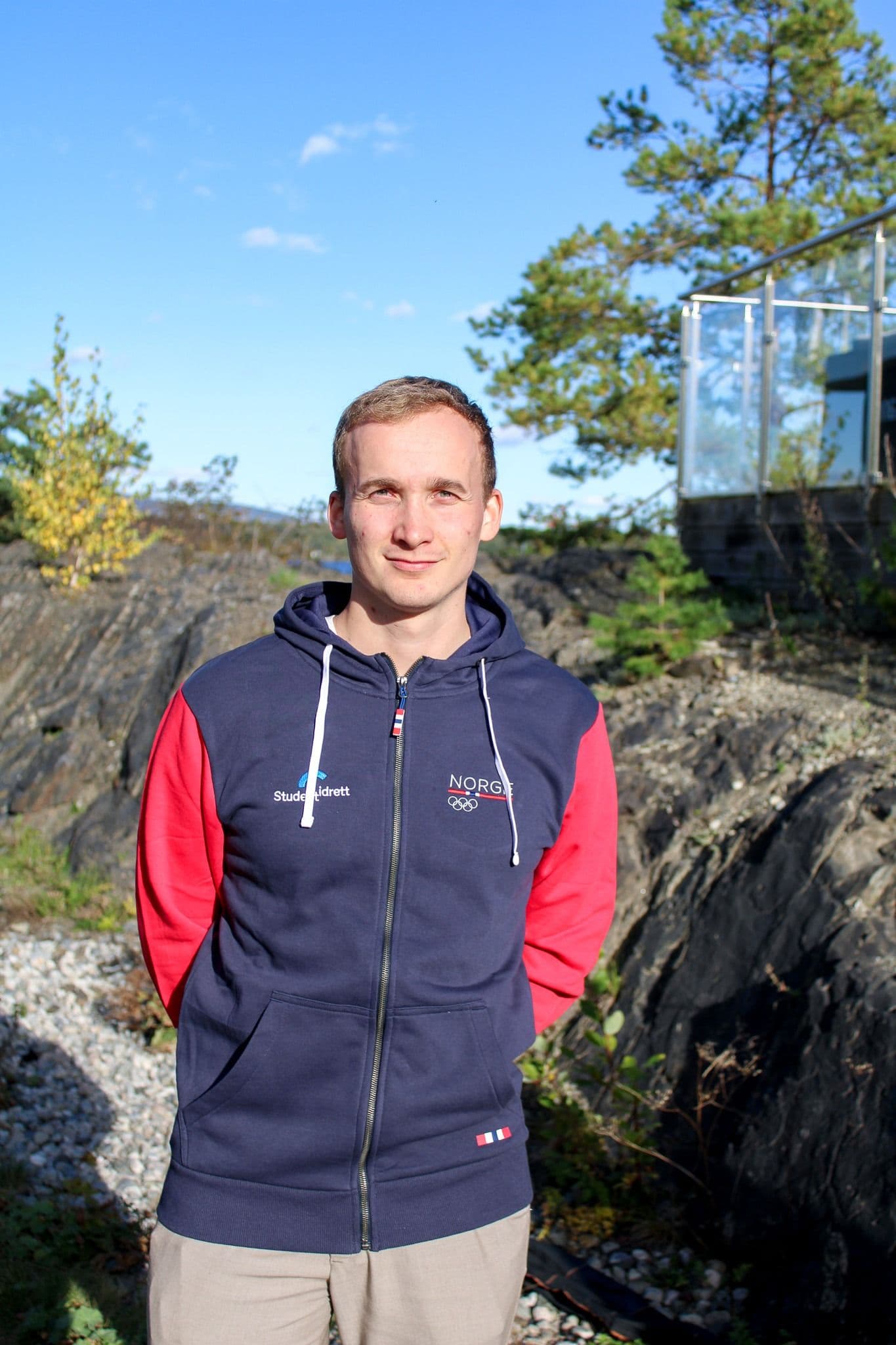 Portrett av en mann i en blå og rød jakke med Studentidrett- og Norges olympiske logoer, fotografert utendørs i et kystlandskap.