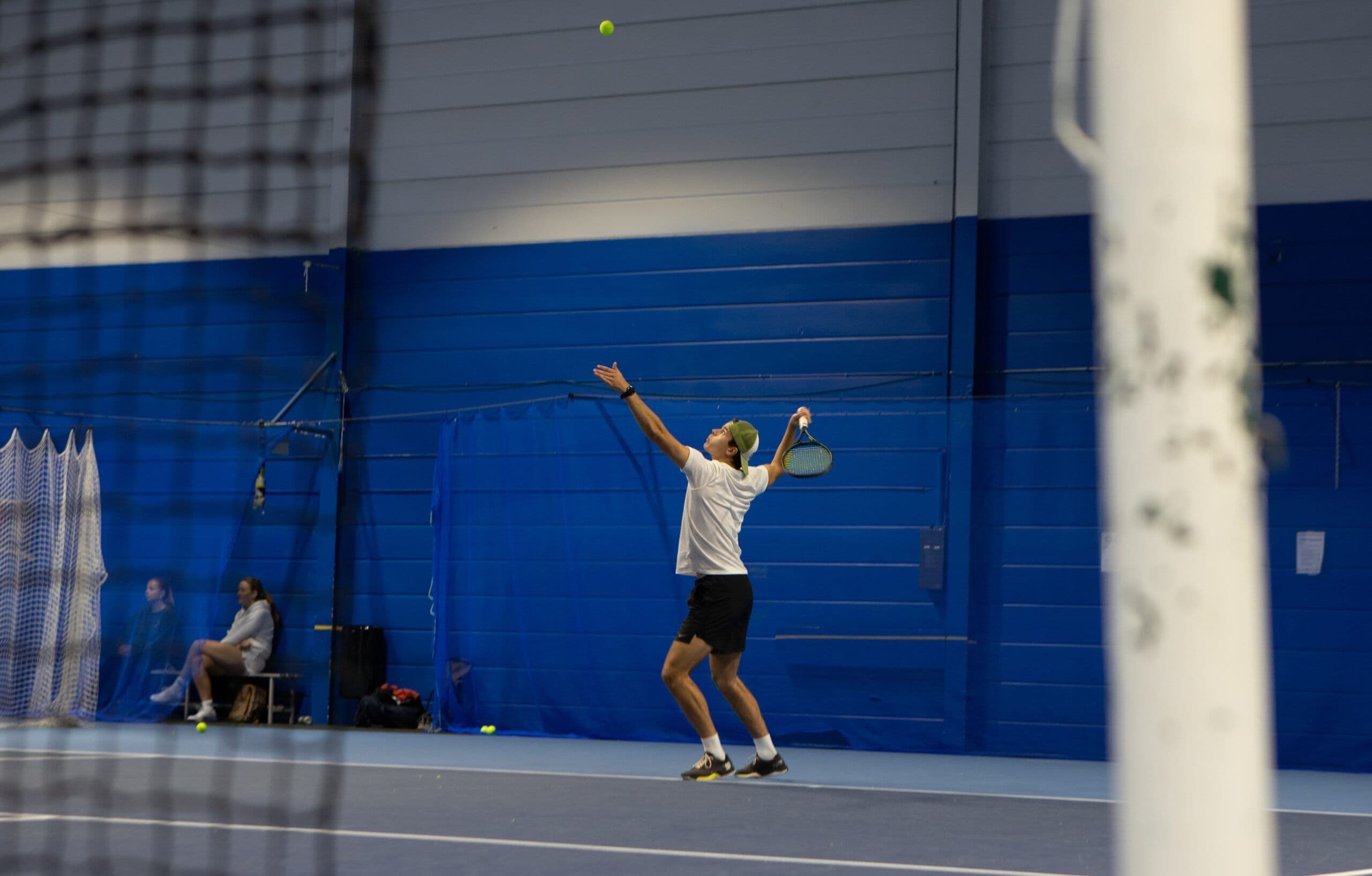 En mannlig tennisspiller strekker seg opp i en serve på en innendørs bane.