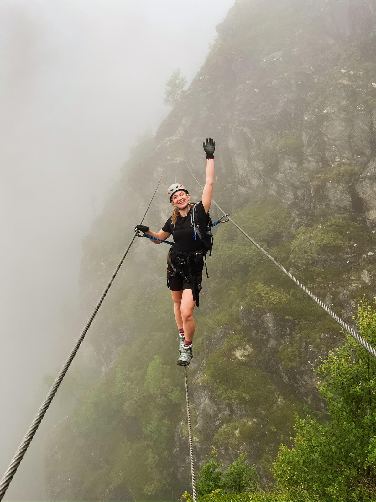 En kvinne med klatrehjelm og sikringsutstyr balanserer på en wire i en via ferrata og strekker den ene armen jublende i været.