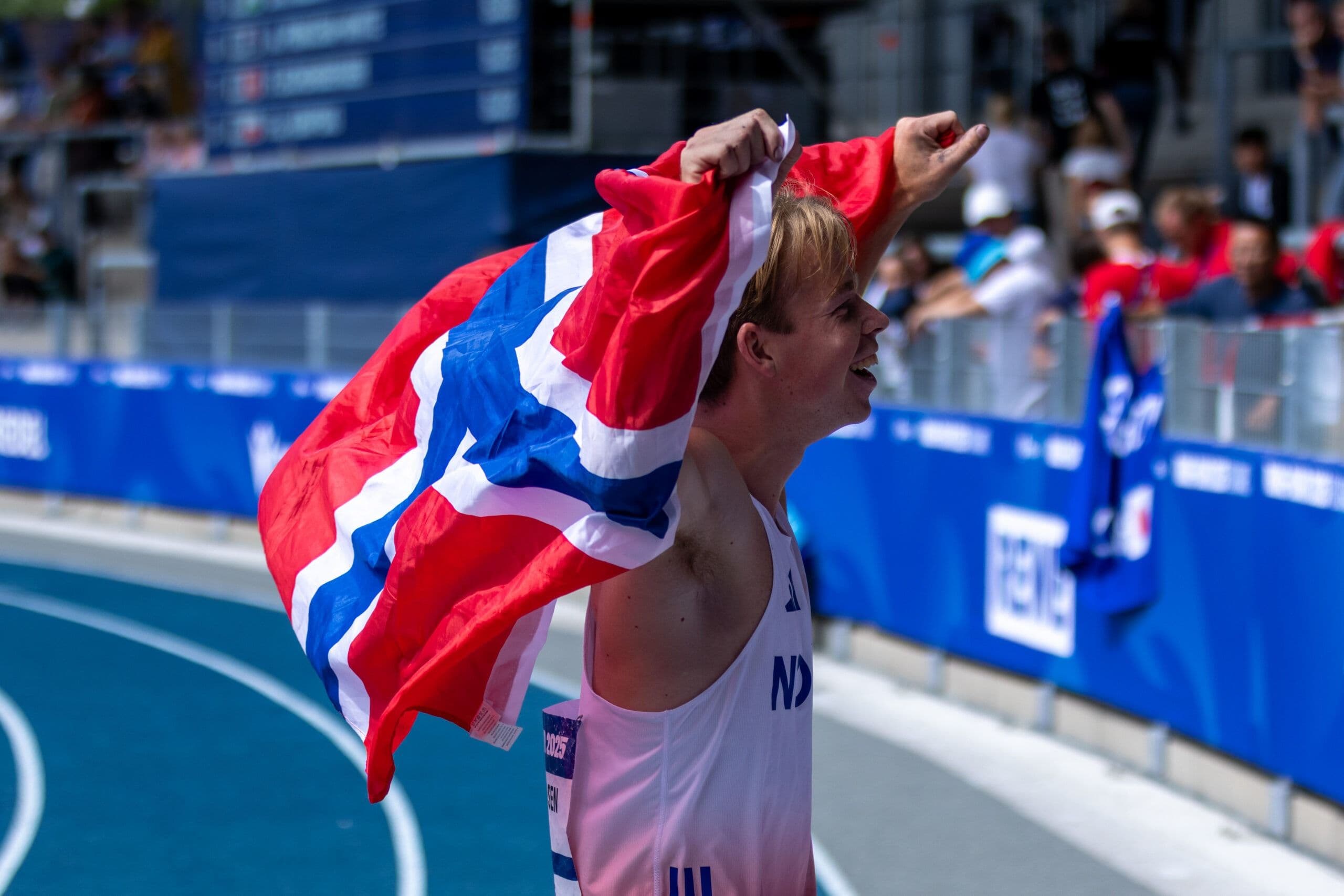 En mannlig friidrettsutøver i Norge-drakt jubler med det norske flagget på løpebanen.