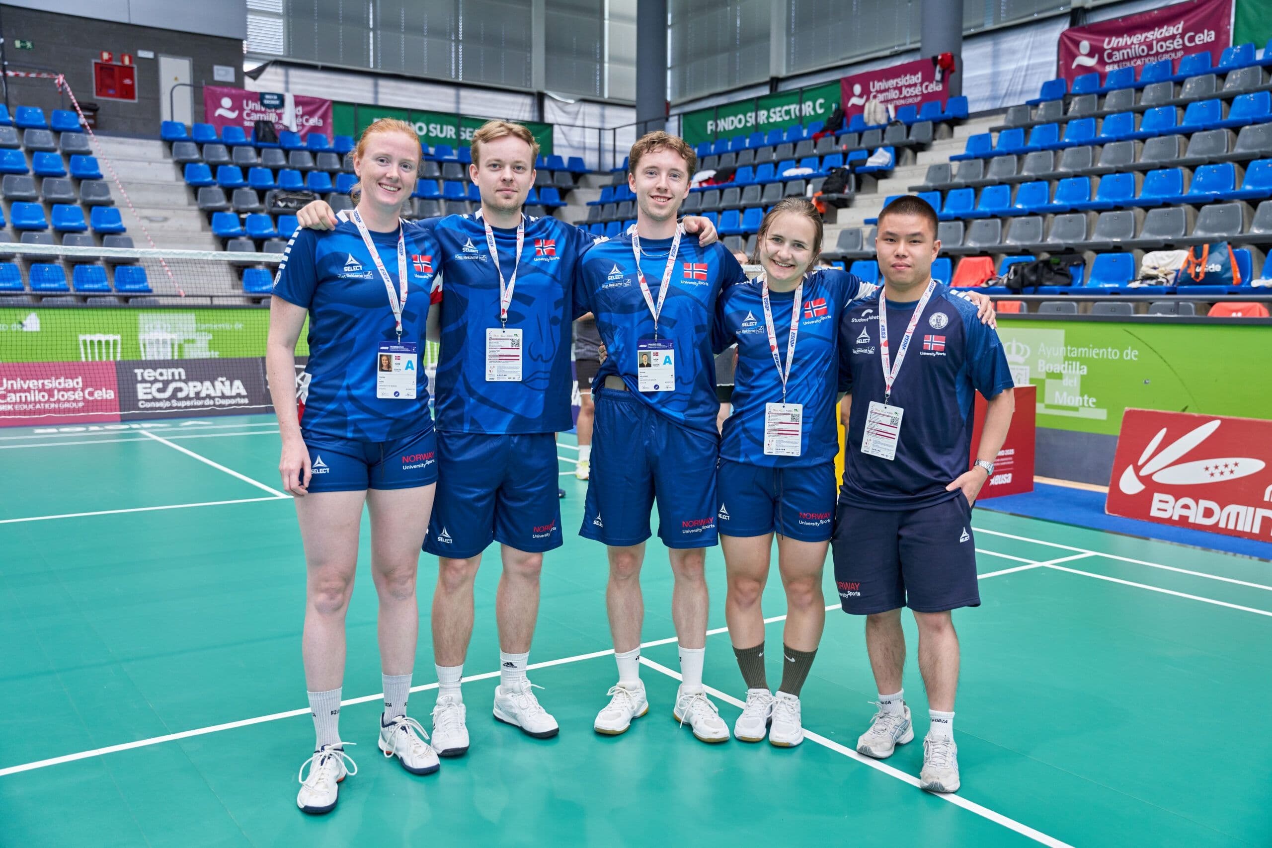 Fem badmintonutøvere i blå landslagsdrakter med akkrediteringskort står samlet på en badmintonbane i en idrettshall.