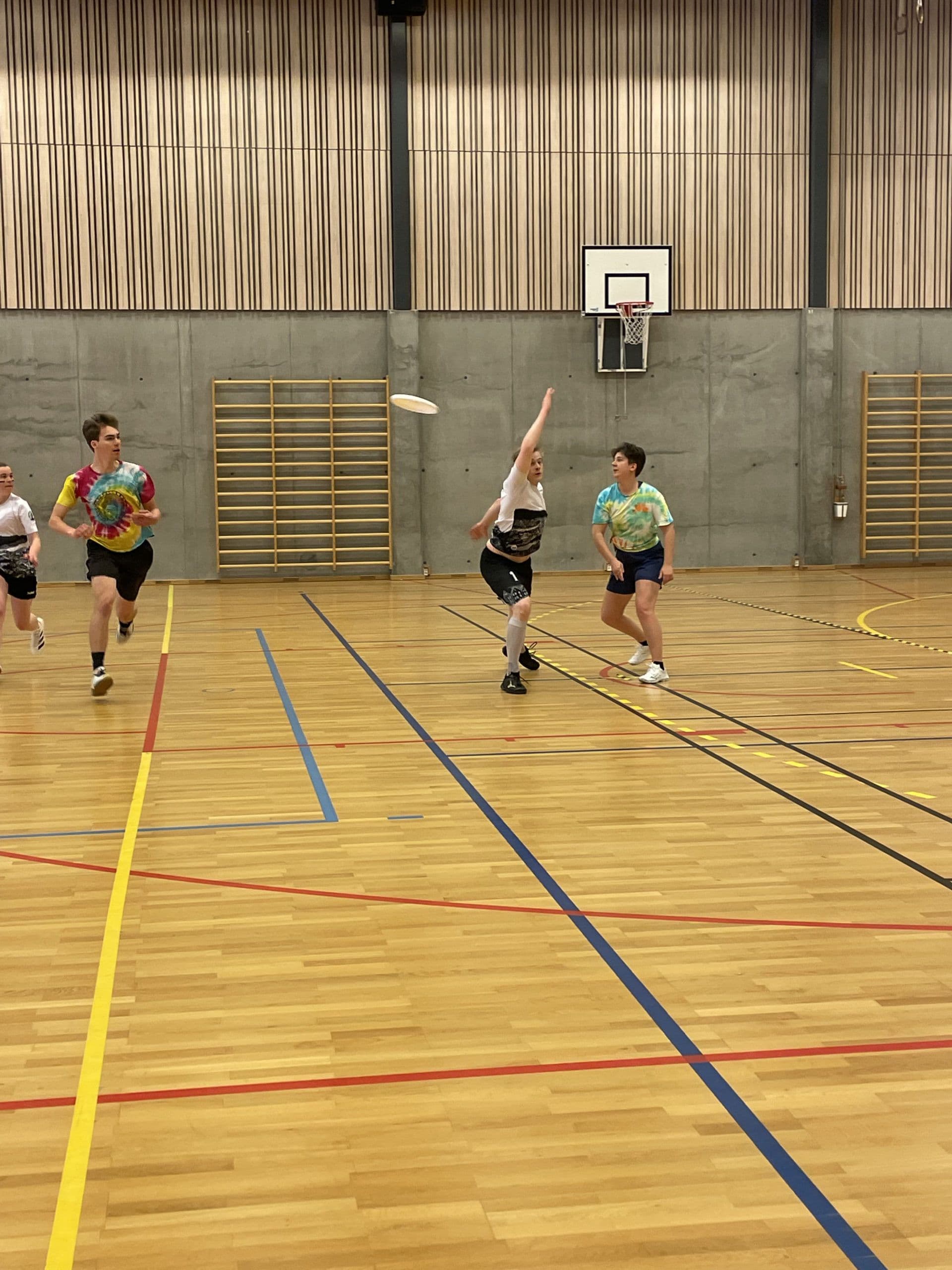 Studenter spiller ultimate frisbee i en idrettshall mens en spiller strekker seg etter frisbeen.