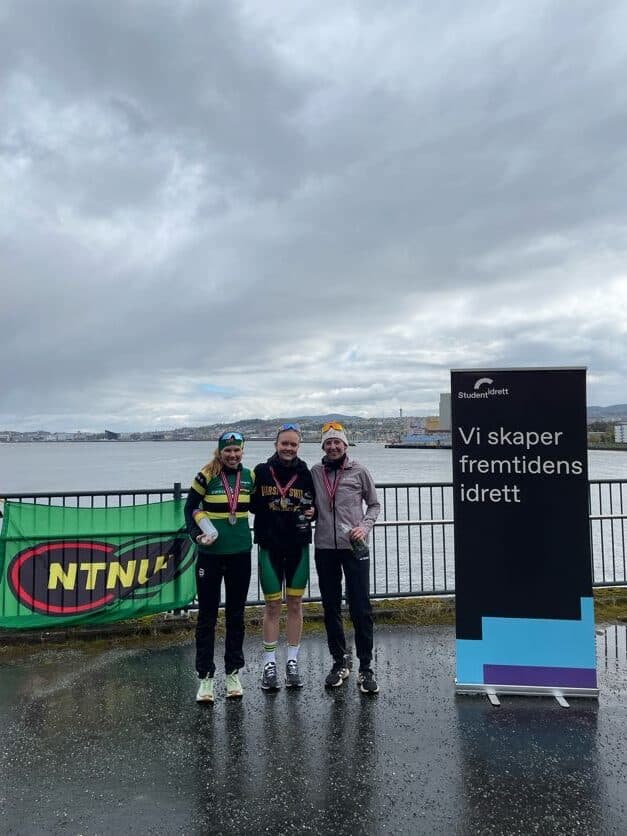 Tre syklister med medaljer om halsen står ved vannet foran et NTNUI-banner og en Studentidrett-rollup med teksten «Vi skaper fremtidens idrett».