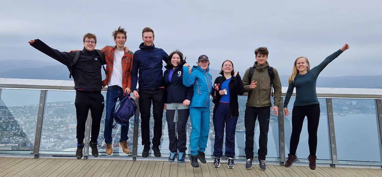 En gruppe studenter poserer med armene i været på en utsiktsplattform med utsikt over hav og by.