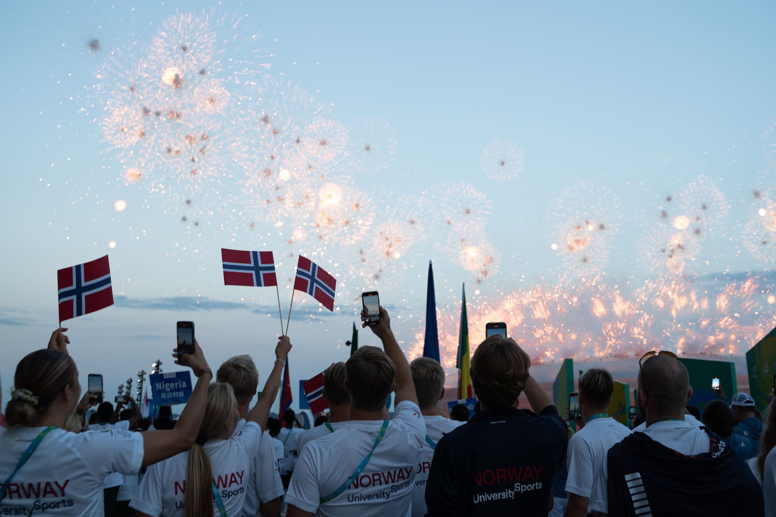 En norsk delegasjon i Norway University Sports-t-skjorter vifter med norske flagg og filmer et fyrverkeri på en åpningsseremoni.