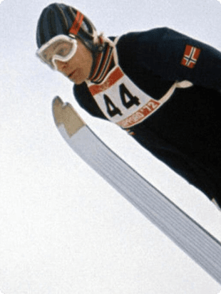 En hoppende skihopper med startnummer 44 og norsk flagg på drakten, fotografert nedenfra mot himmelen.
