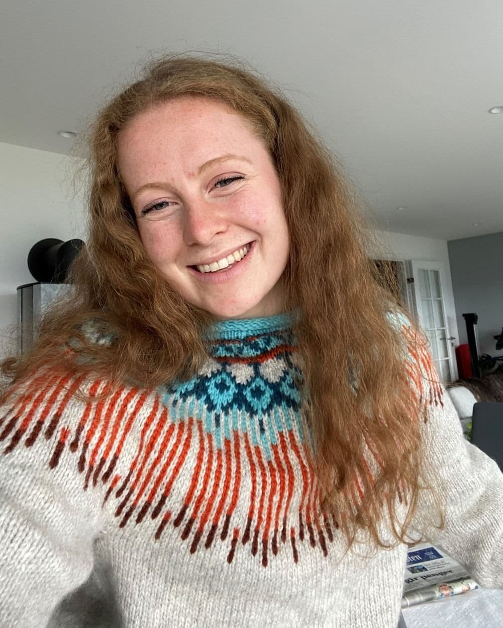 Selfieportrett av en smilende kvinne med rødt krøllete hår i en mønstret ullgenser innendørs.