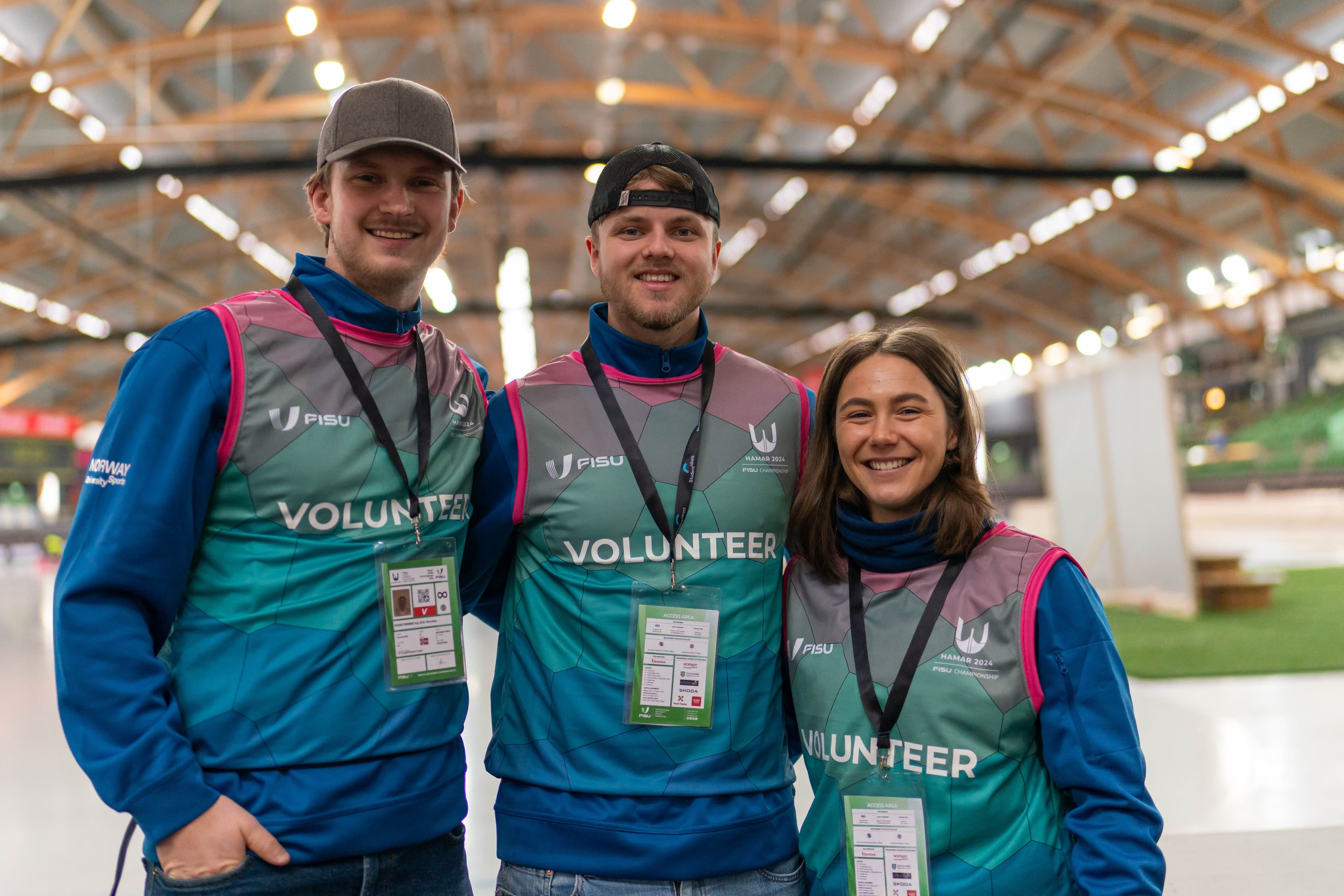 Tre frivillige i turkise «Volunteer»-vester smiler i en skøytehall under FISU World University Games.