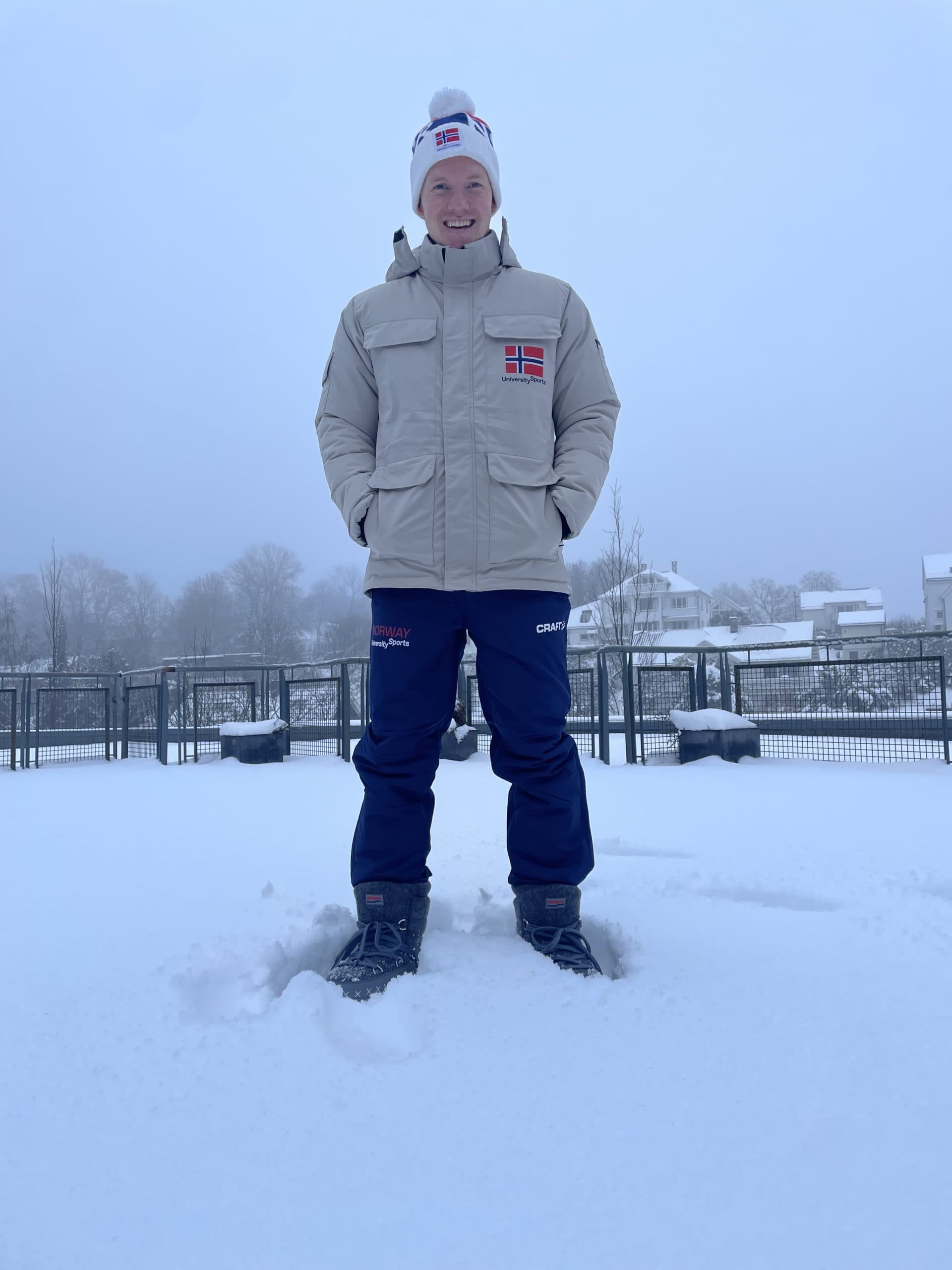 En person i norsk landslagsantrekk med lue og vinterjakke står smilende i dyp snø.