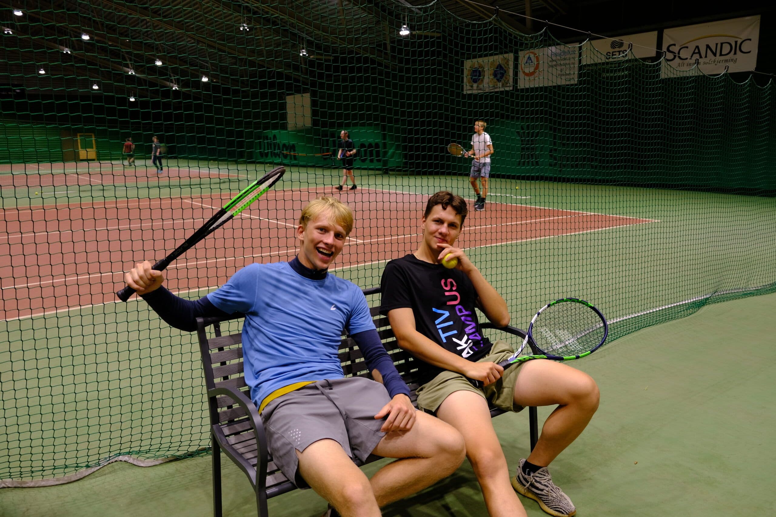 To unge menn med tennisracketer sitter på en benk ved en innendørs tennisbane.