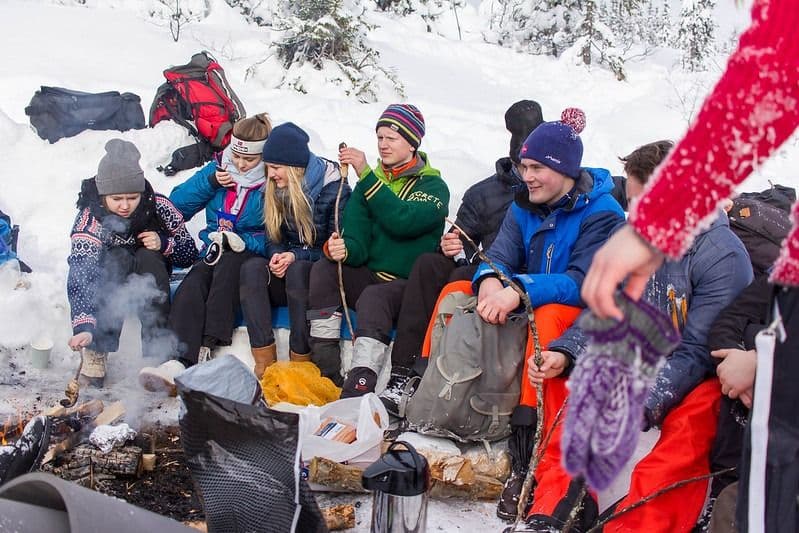 En gruppe unge mennesker i vinterklær sitter samlet rundt et bål i snøen og griller.
