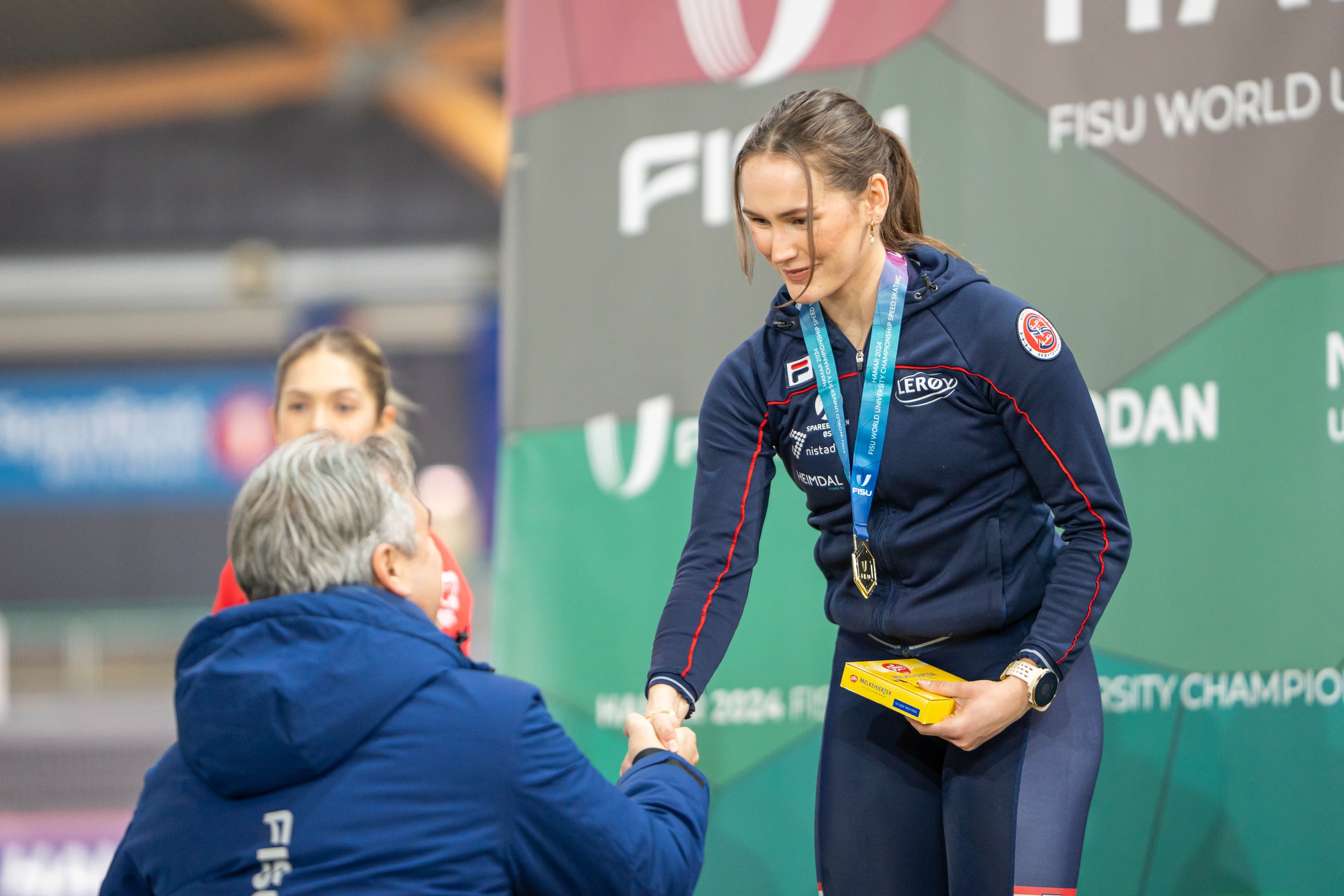 En kvinnelig skøyteløper i norsk landslagsjakke tar imot medalje og håndhilser på premieutdelingen under et FISU-mesterskap.